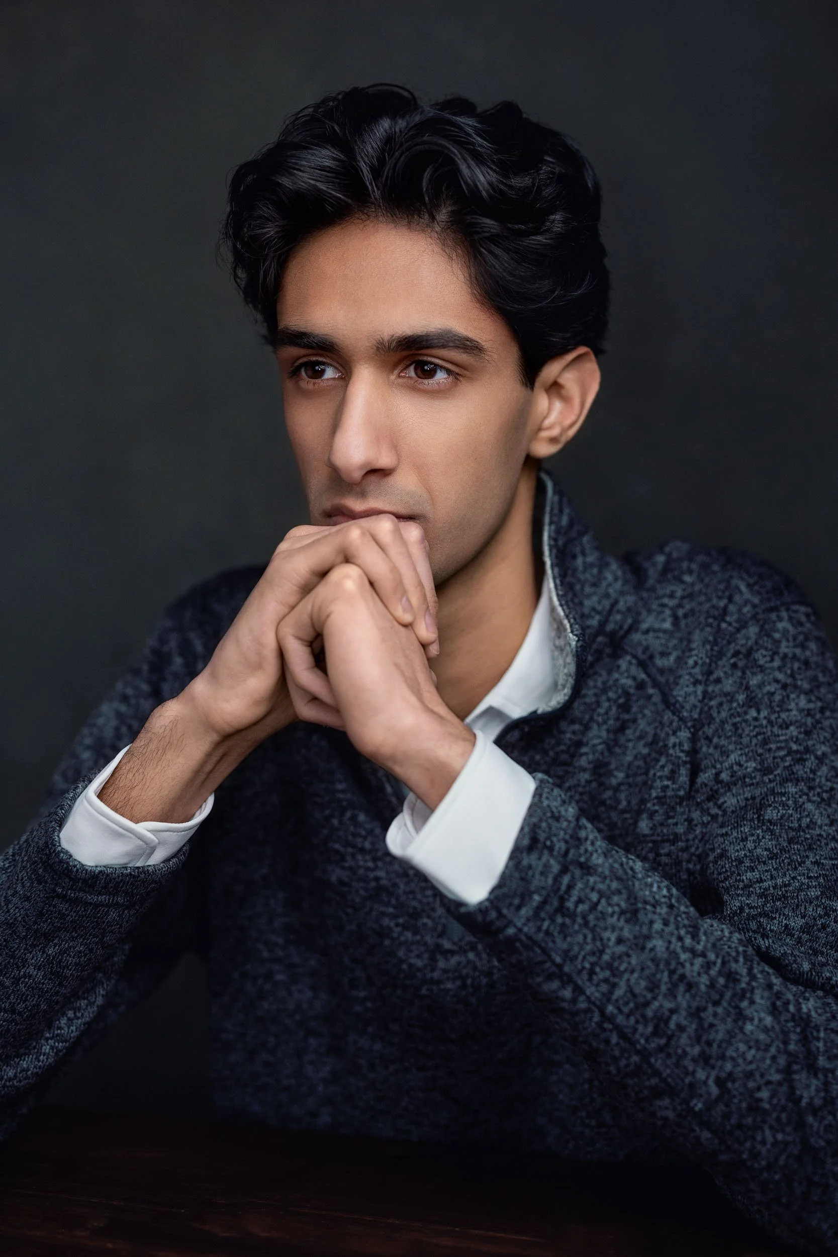 A young man with dark, wavy hair and light brown skin sitting with his hands clasped near his mouth, wearing a dark gray sweater over a white shirt, against a dark background.