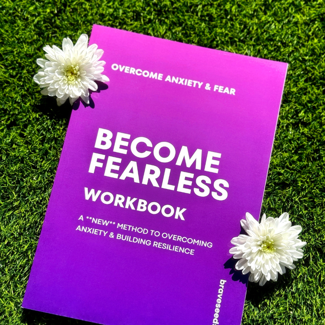 A purple workbook titled 'Become Fearless Workbook' with white flowers beside it, placed on green grass.
