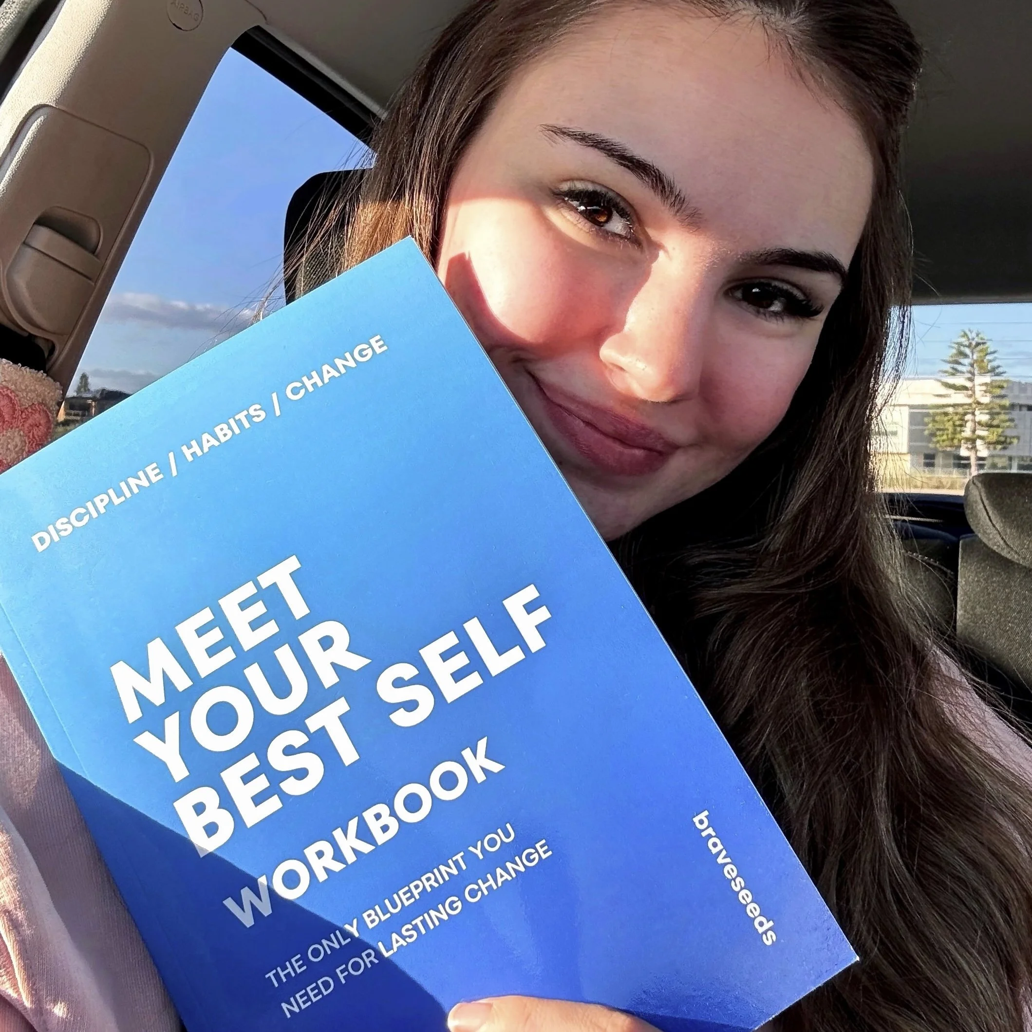 A young woman with long brown hair holding a blue booklet titled 'FIFTY TIPS / HABITS / CHANGE,' smiling inside a vehicle with a view of blue sky and trees outside.