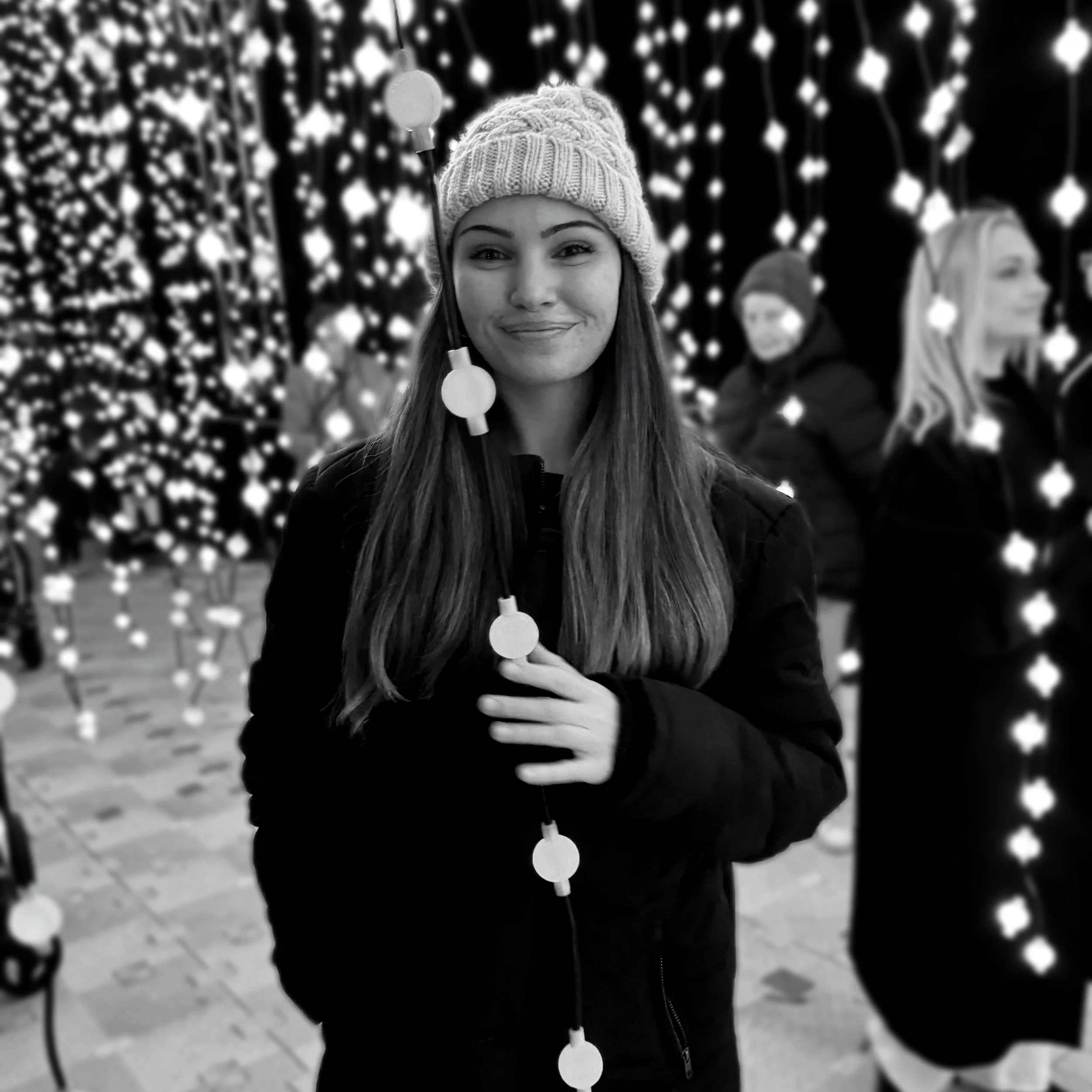 A woman wearing a knit hat and a dark jacket, smiling at the camera, holding a string of round lights, with a background of many small round lights hanging and decorations, at a festive event or holiday display.