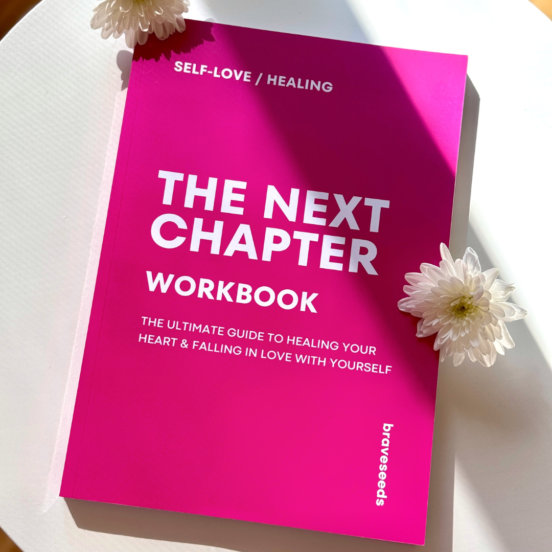 Pink workbook titled 'The Next Chapter Workbook' with white flowers on top, placed on a white surface, emphasizing self-love and healing.