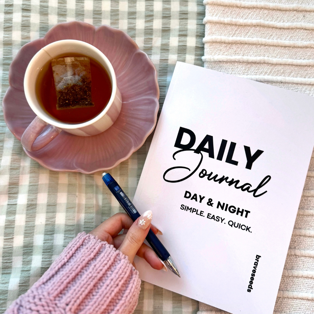A pink plate with a tea cup on top, a person's hand holding a pen, a journal titled "Daily Journal" with the subtitle "Day & Night" and the words "Simple, Easy, Quick." on a checkered cloth.