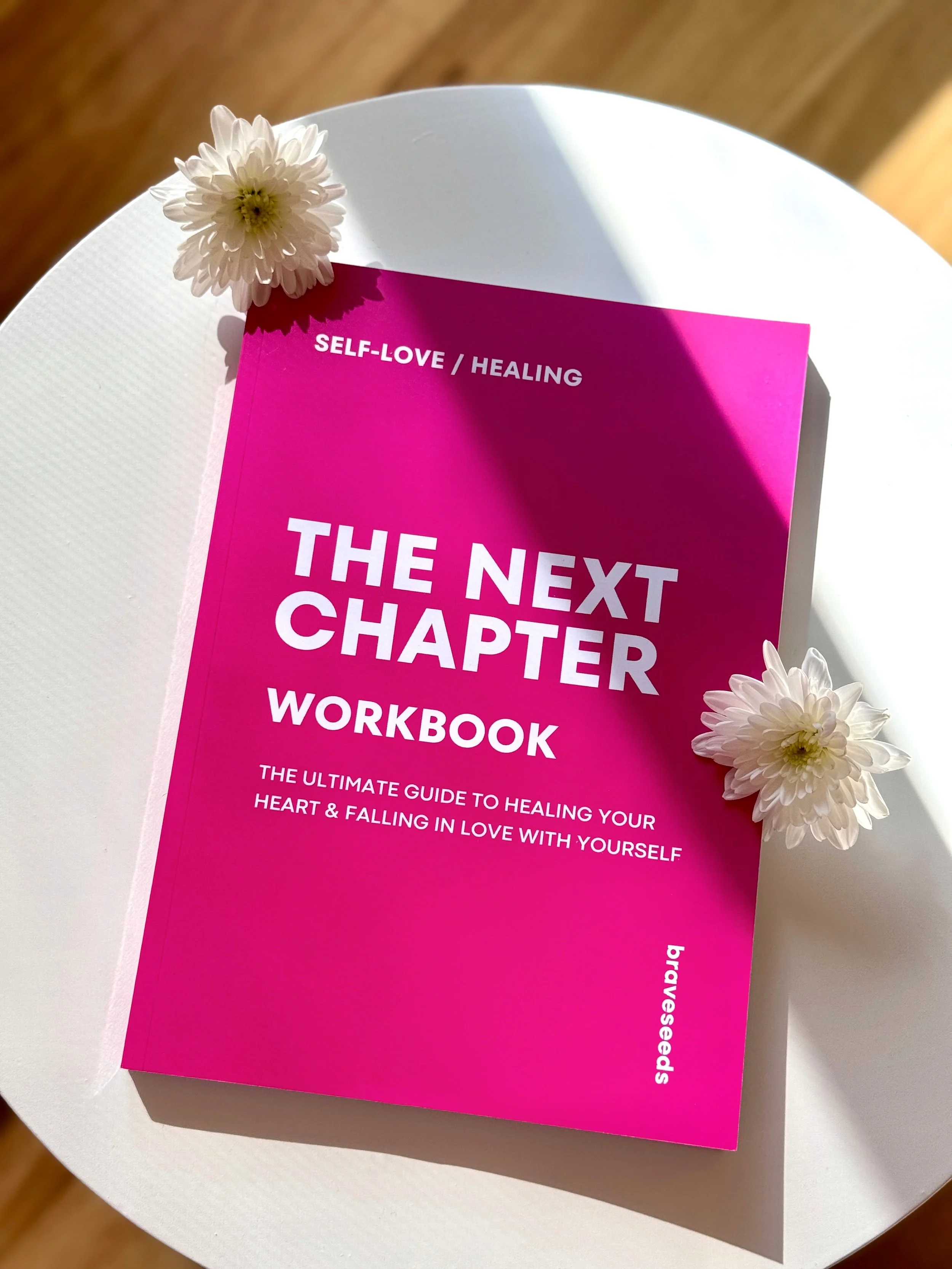 A pink workbook titled "The Next Chapter Workbook" with white text, placed on a white table with white flowers and natural sunlight shining on it.