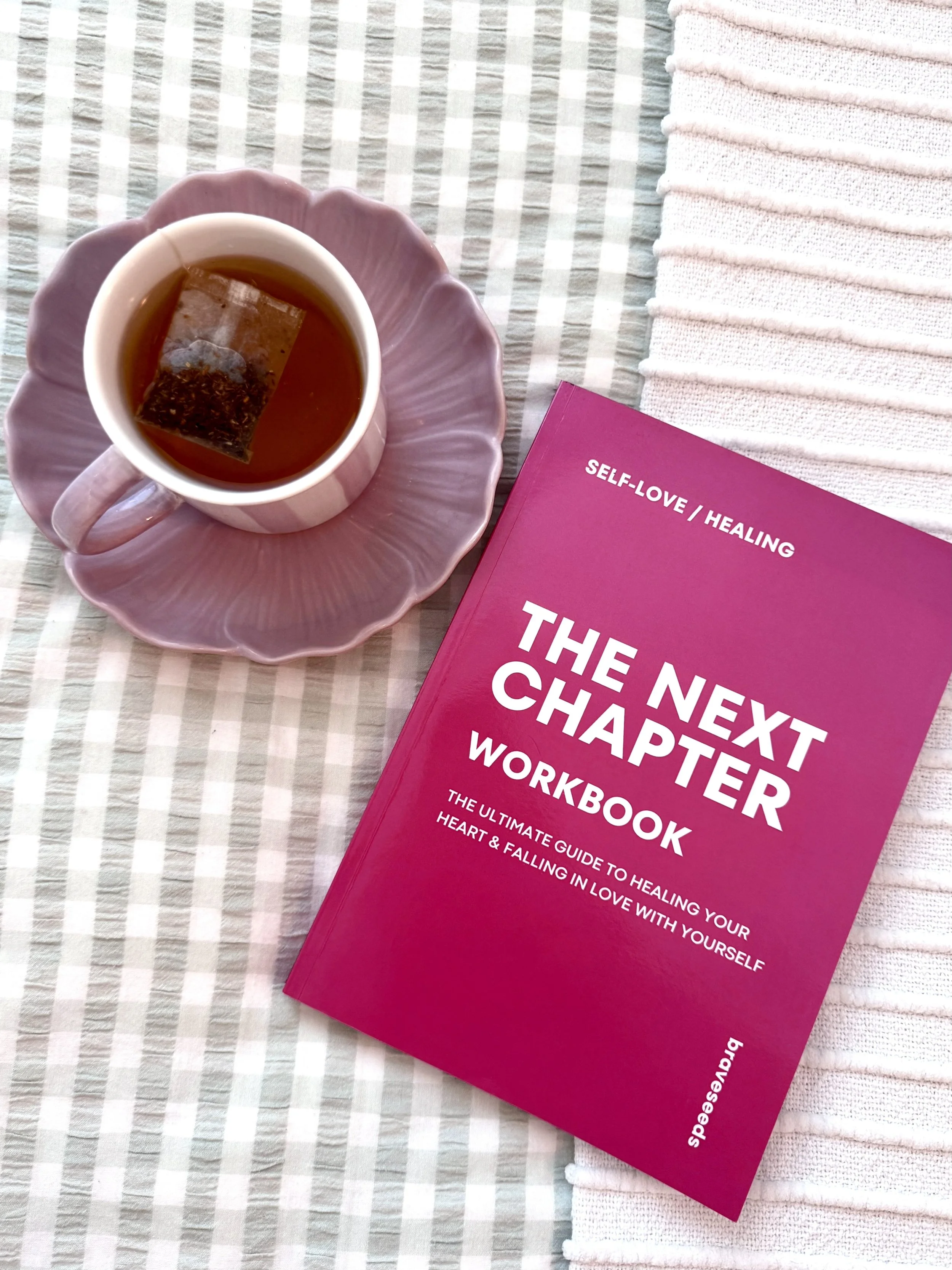 A pink book titled 'The Next Chapter Workbook' with the subtitle 'Self-Love / Healing' on a striped tablecloth, next to a pink scalloped dish with a white teacup filled with tea, and a tea bag inside.