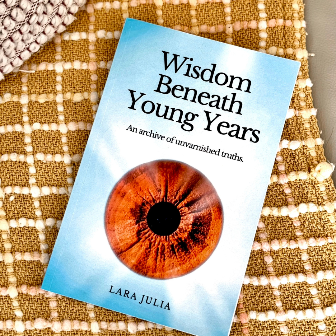 A book titled "Wisdom Beneath Young Years" by Lara Julia, with a subtitle "An archive of unvarnished truths," lies on a textured, checkered fabric surface. The cover features a close-up image of a human eye with a reddish-brown iris.