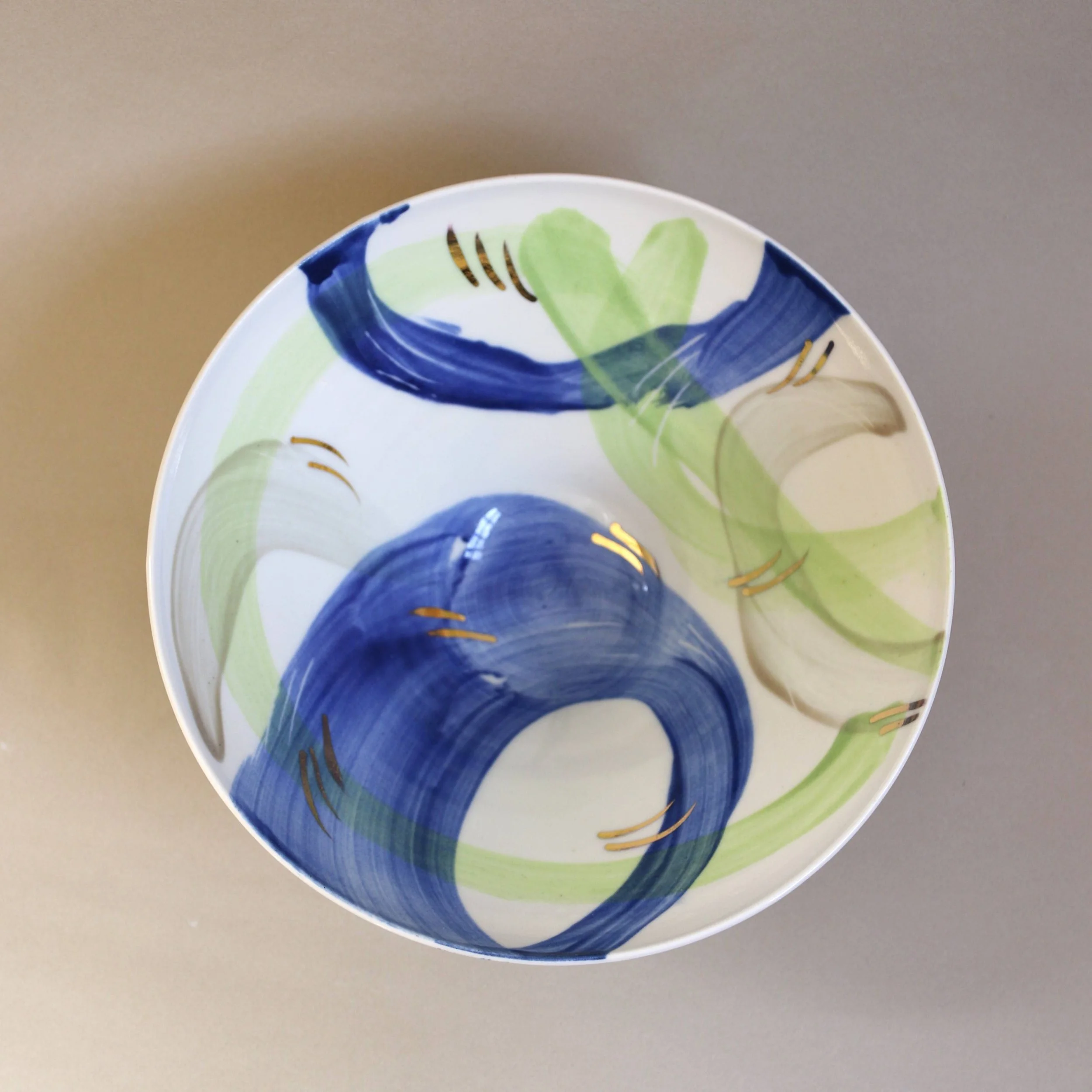 Close-up of a colorful ceramic plate with abstract green, blue, and gold painted design, shown from above against a gray background.