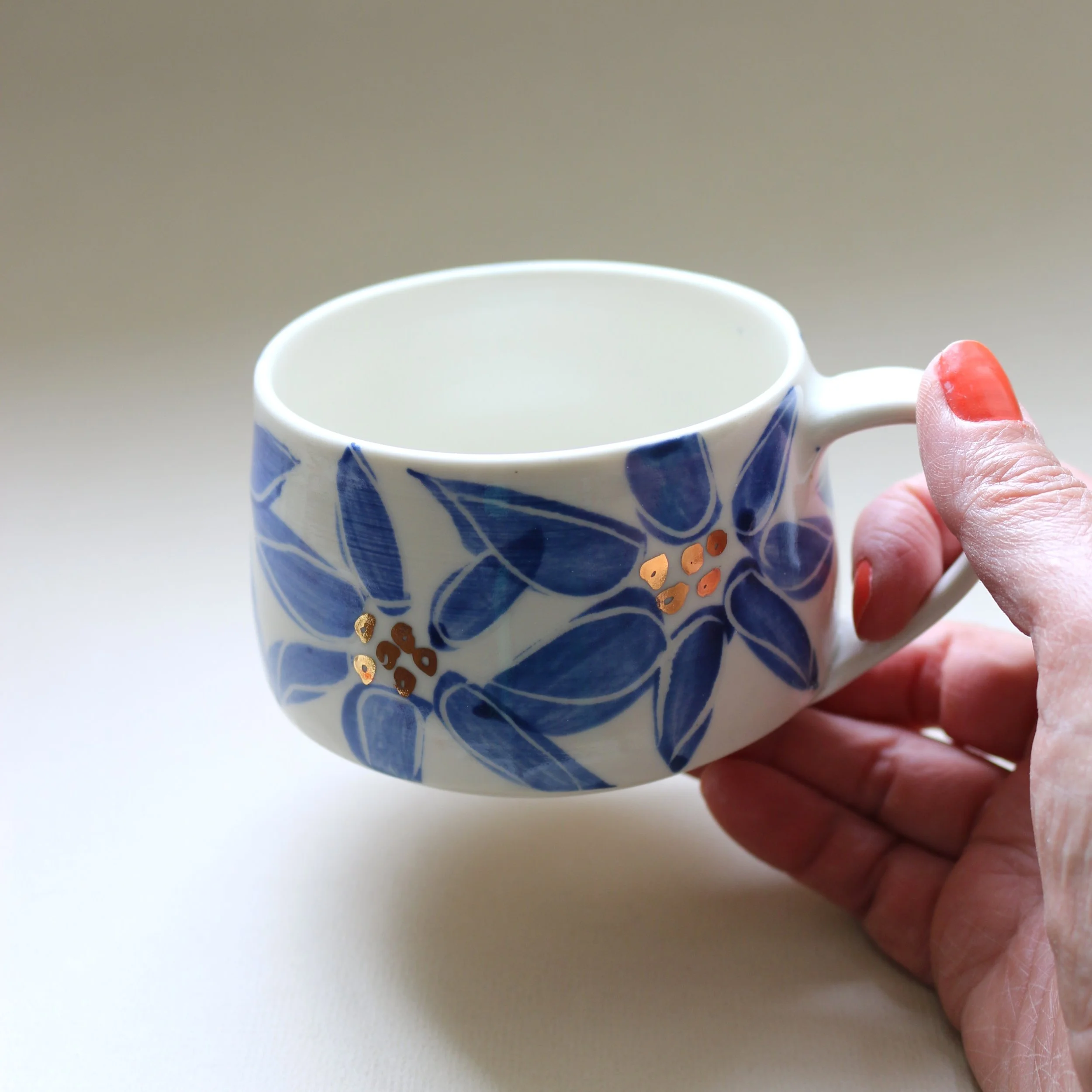 A hand with red-painted nails holding a blue and white patterned ceramic mug on a white surface.