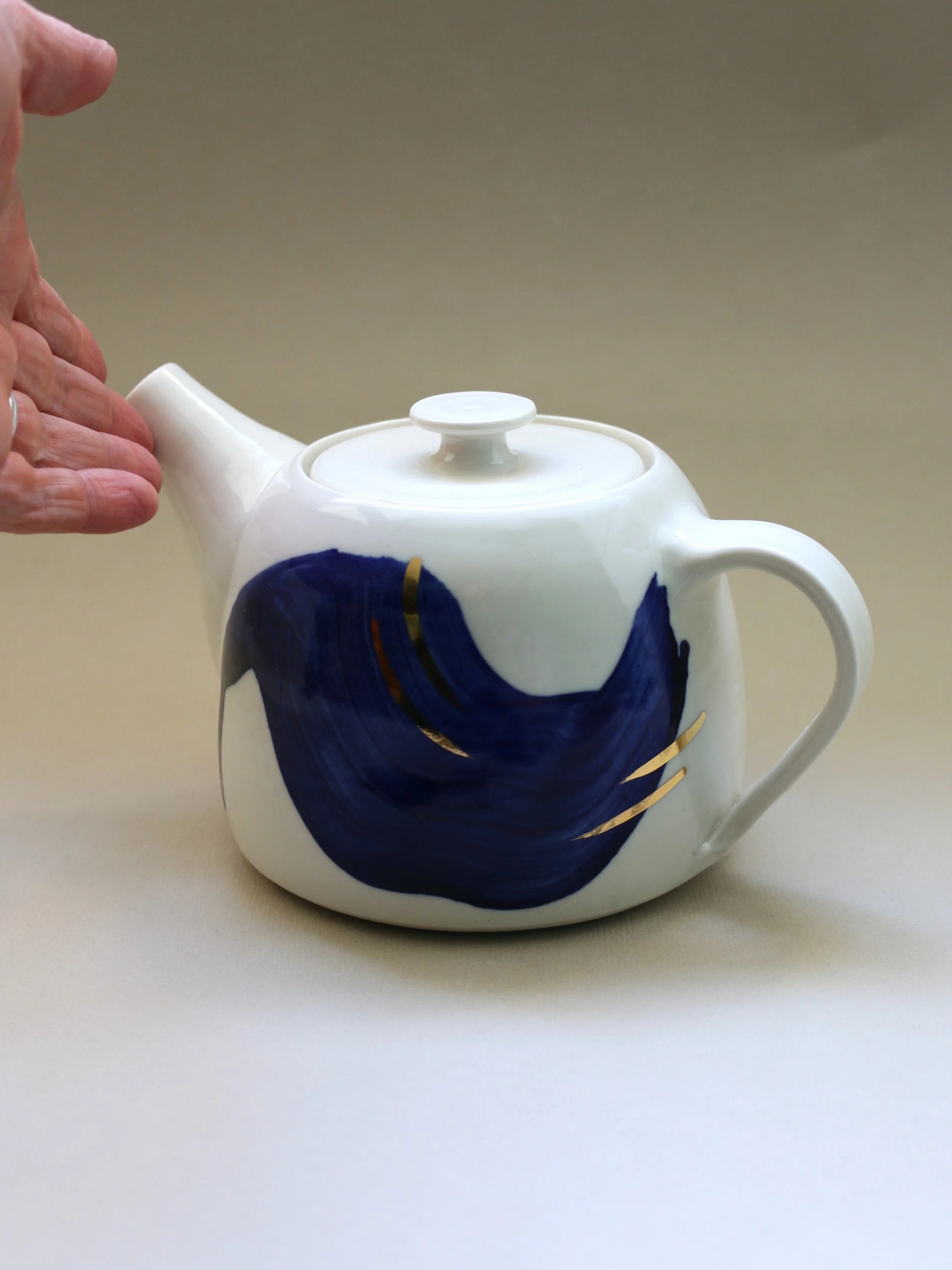 A white ceramic teapot with a blue abstract design on its body, topped with a white lid, on a cream-colored surface.