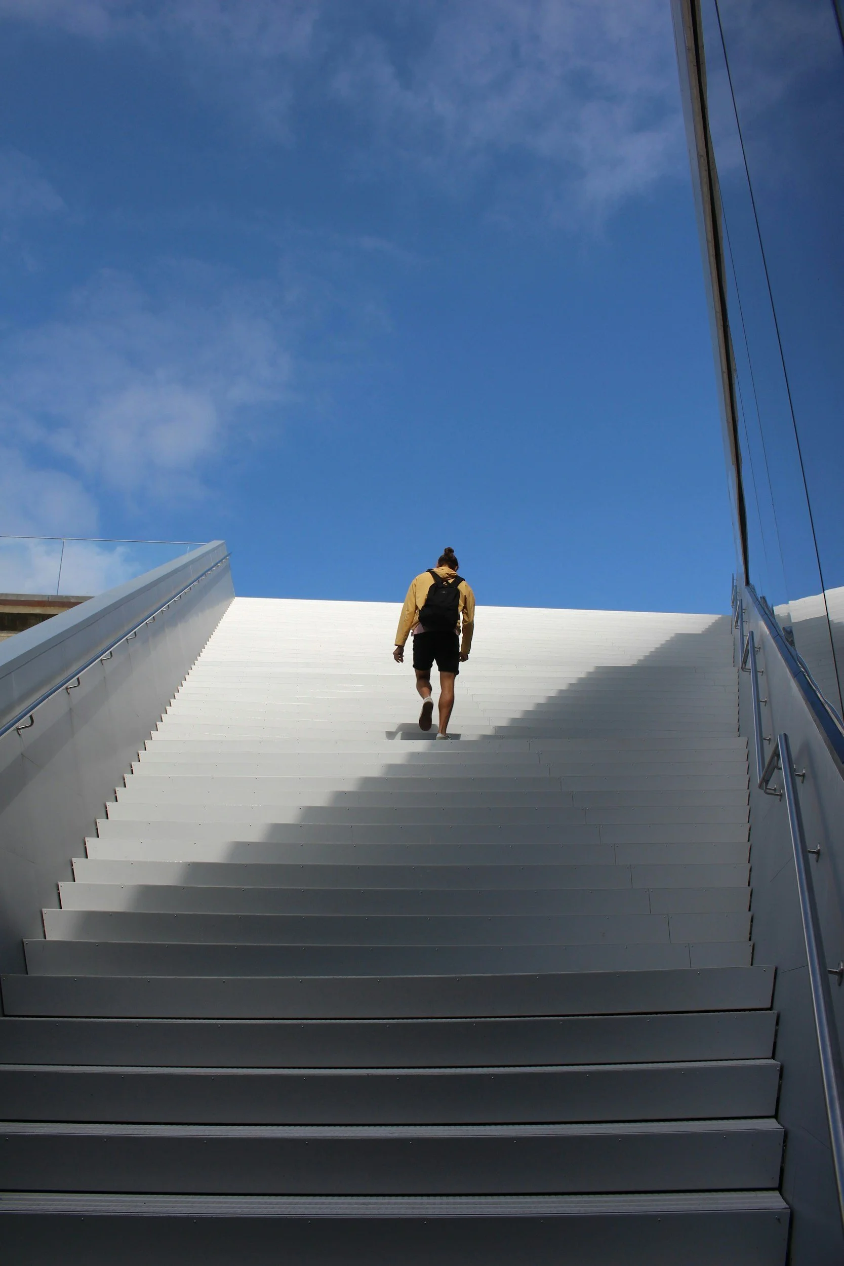 Person with a backpack ascending white stairs outside, blue sky with some clouds above, modern building with reflective glass windows on the right side.