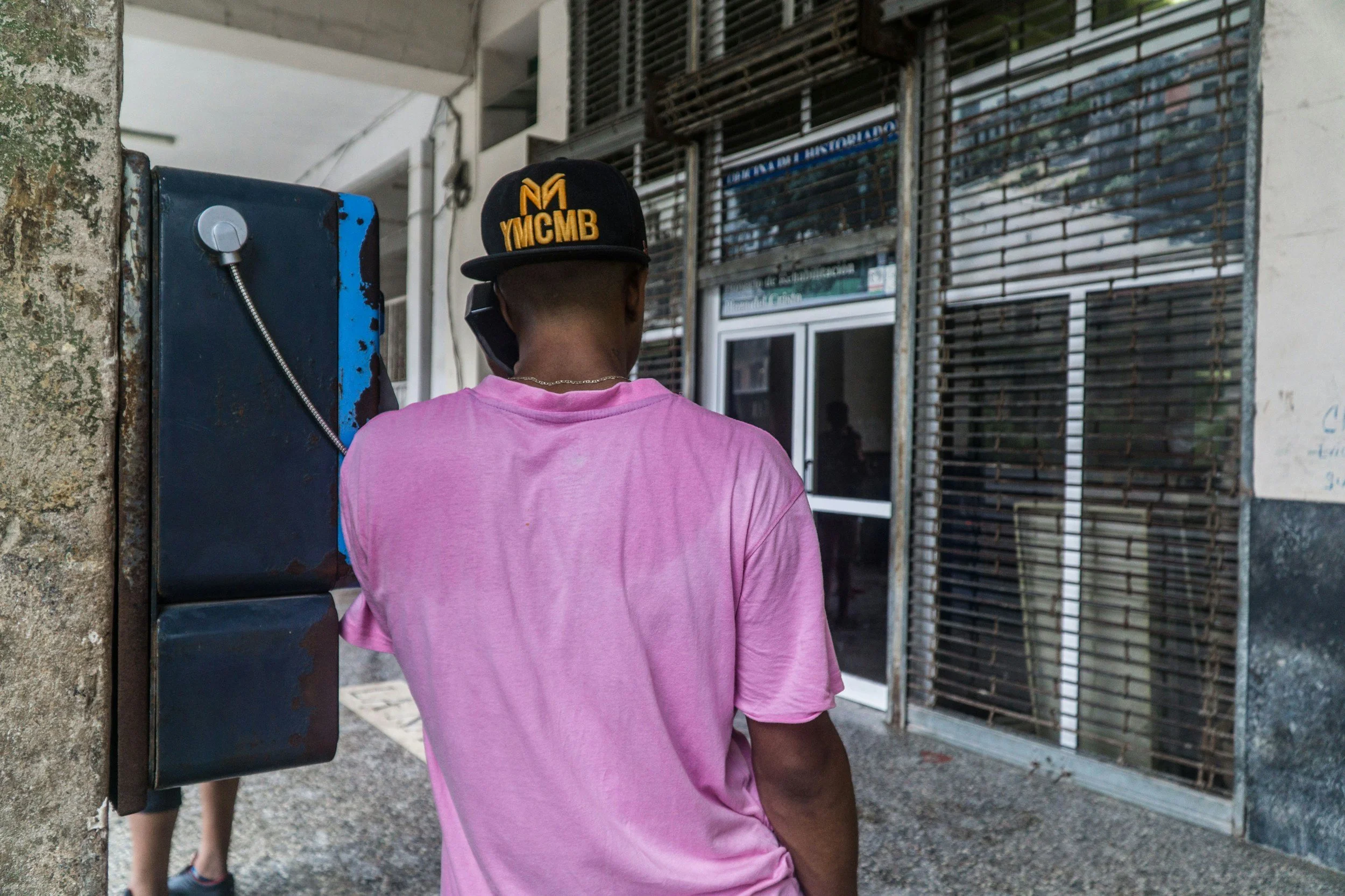 Person wearing a black cap with yellow lettering, pink T-shirt, talking on a mobile phone, near an outdoor phone booth, with a building with metal grills in the background.