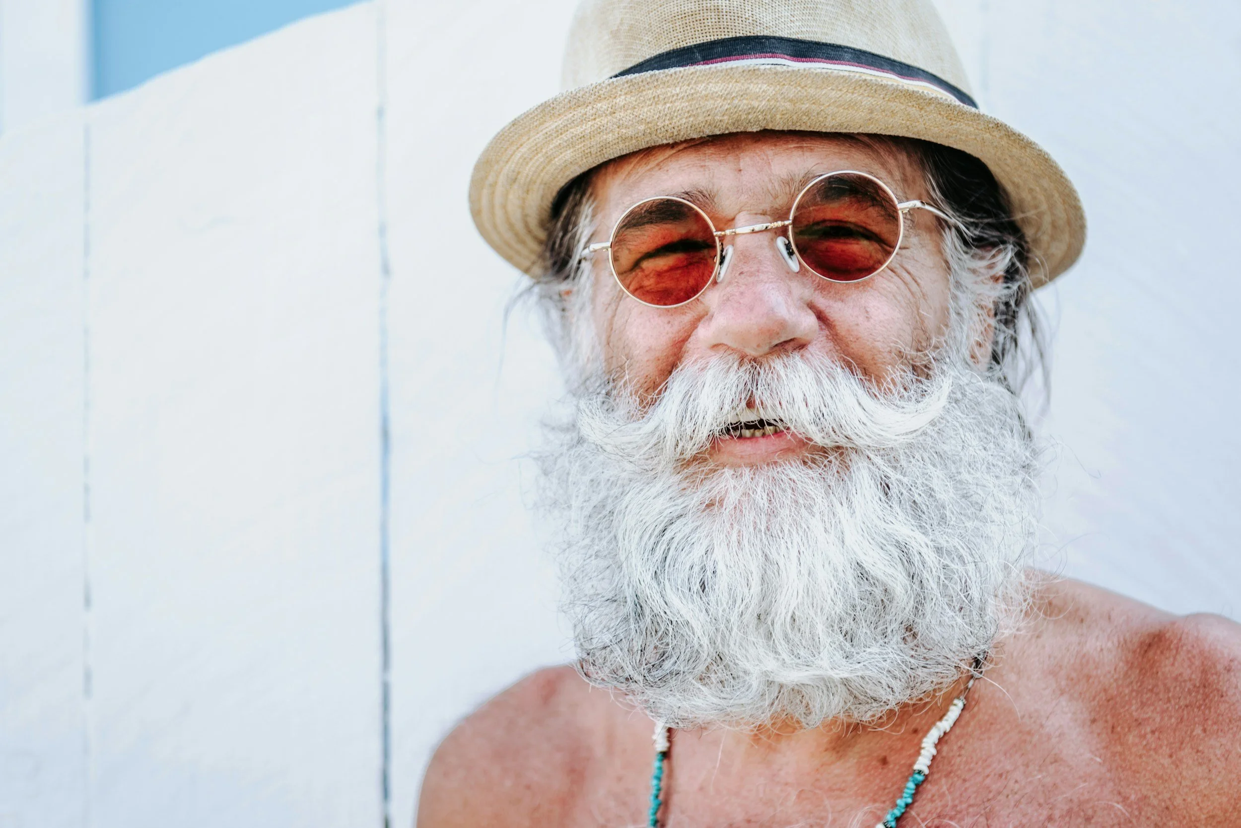 Close-up of smiling older man with a beard and mustache wearing sunglasses, a straw hat, and necklaces, against a white wall background.