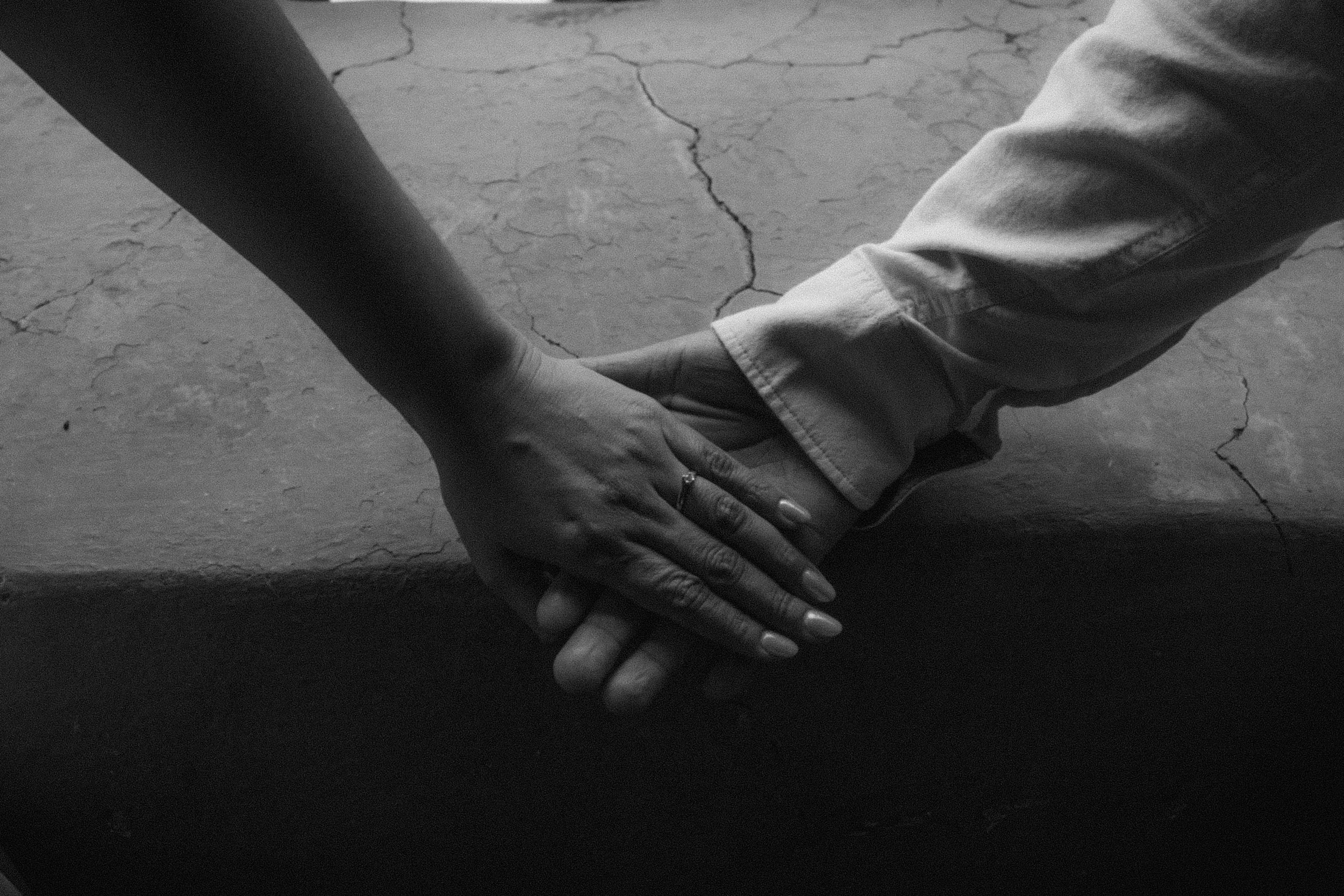 Close-up of two hands holding each other, with one person wearing a ring. The background shows a cracked concrete floor.