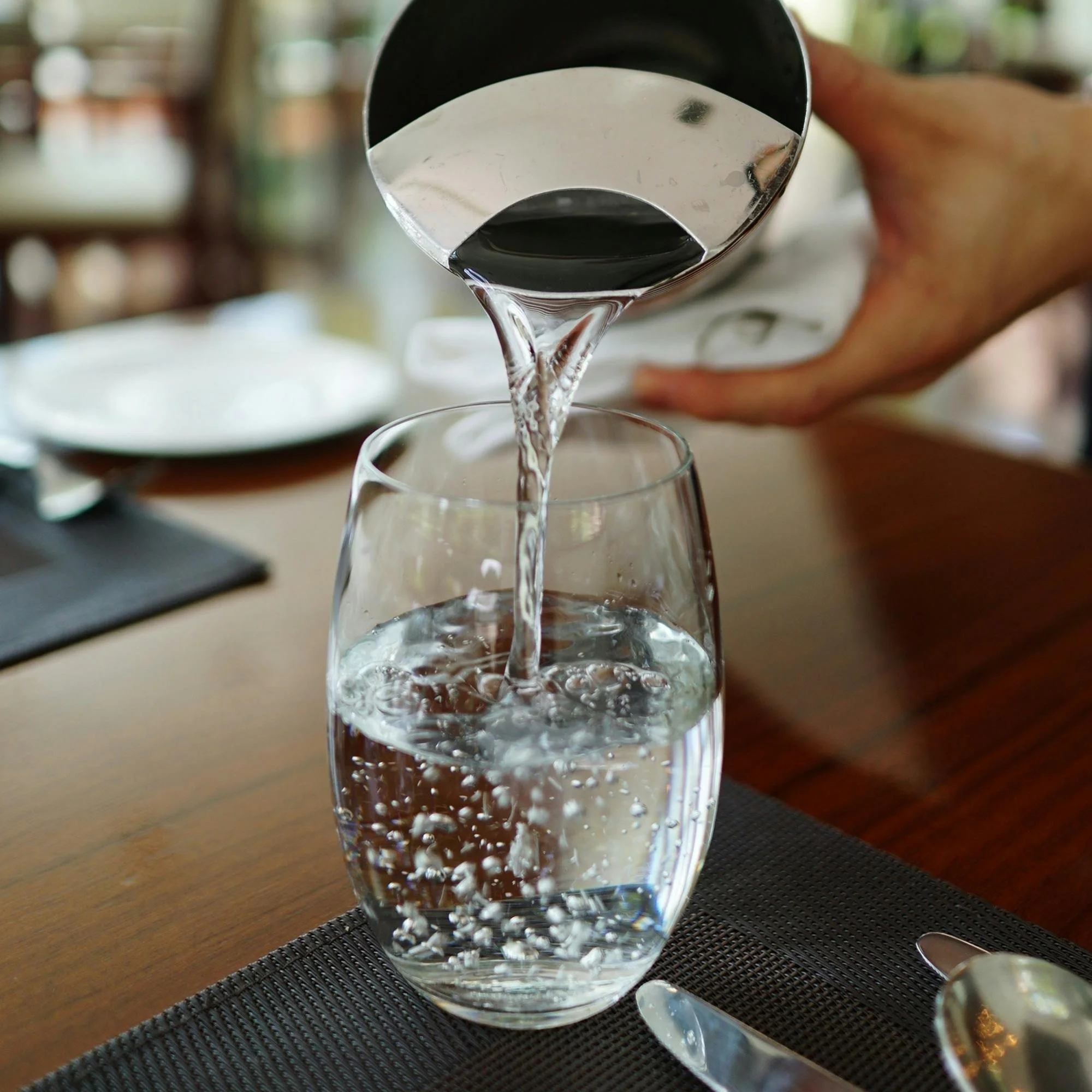 A person pouring water from a bottle into a clear glass. The scene is set on a wooden table with a black placemat, and the background is out of focus.