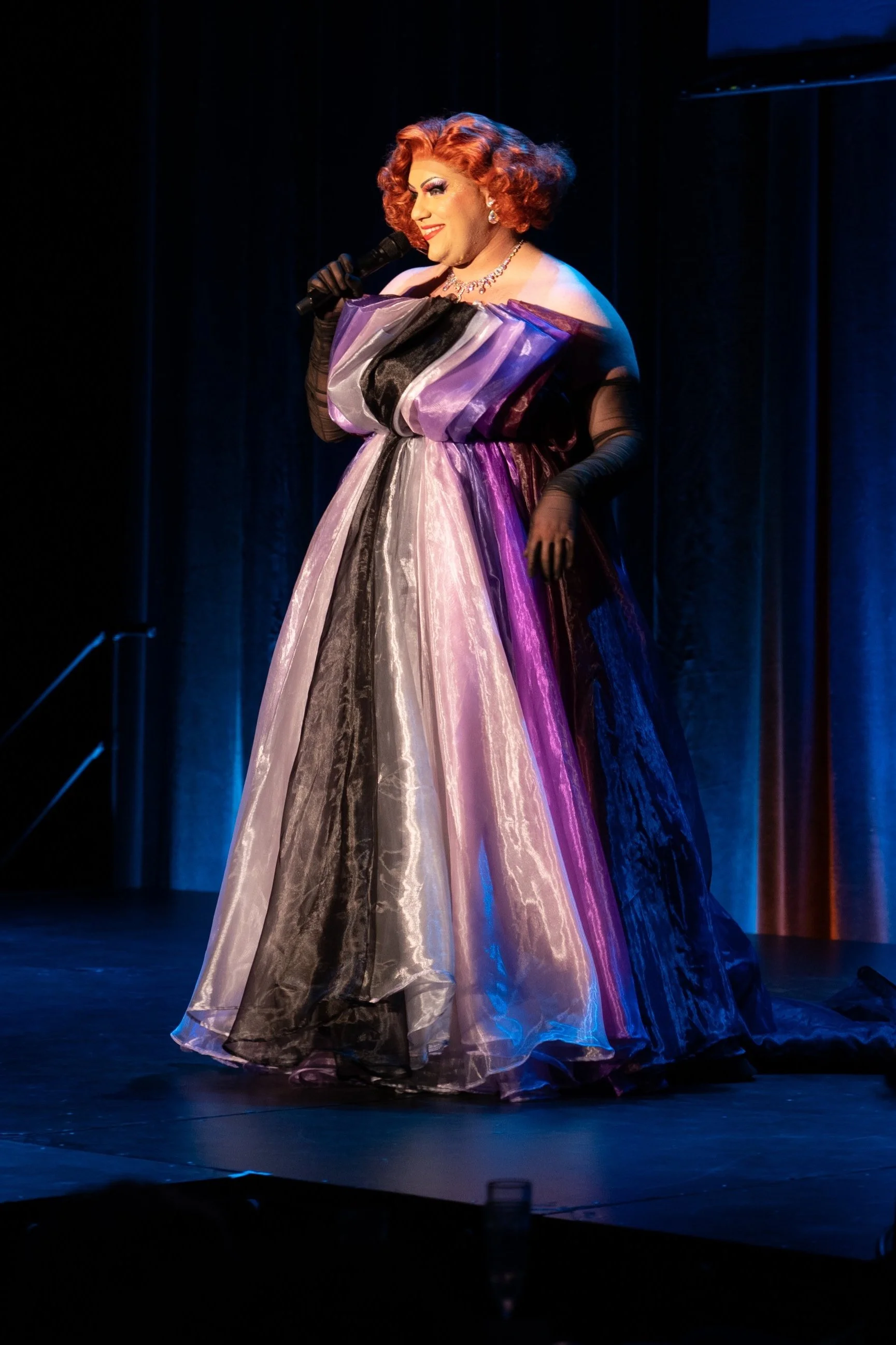 A performer on stage dressed in a colorful, shiny gown with a voluminous skirt, black gloves, and dramatic makeup, holding a microphone. Broadway Divas SCFTA Miss Clair Voyance