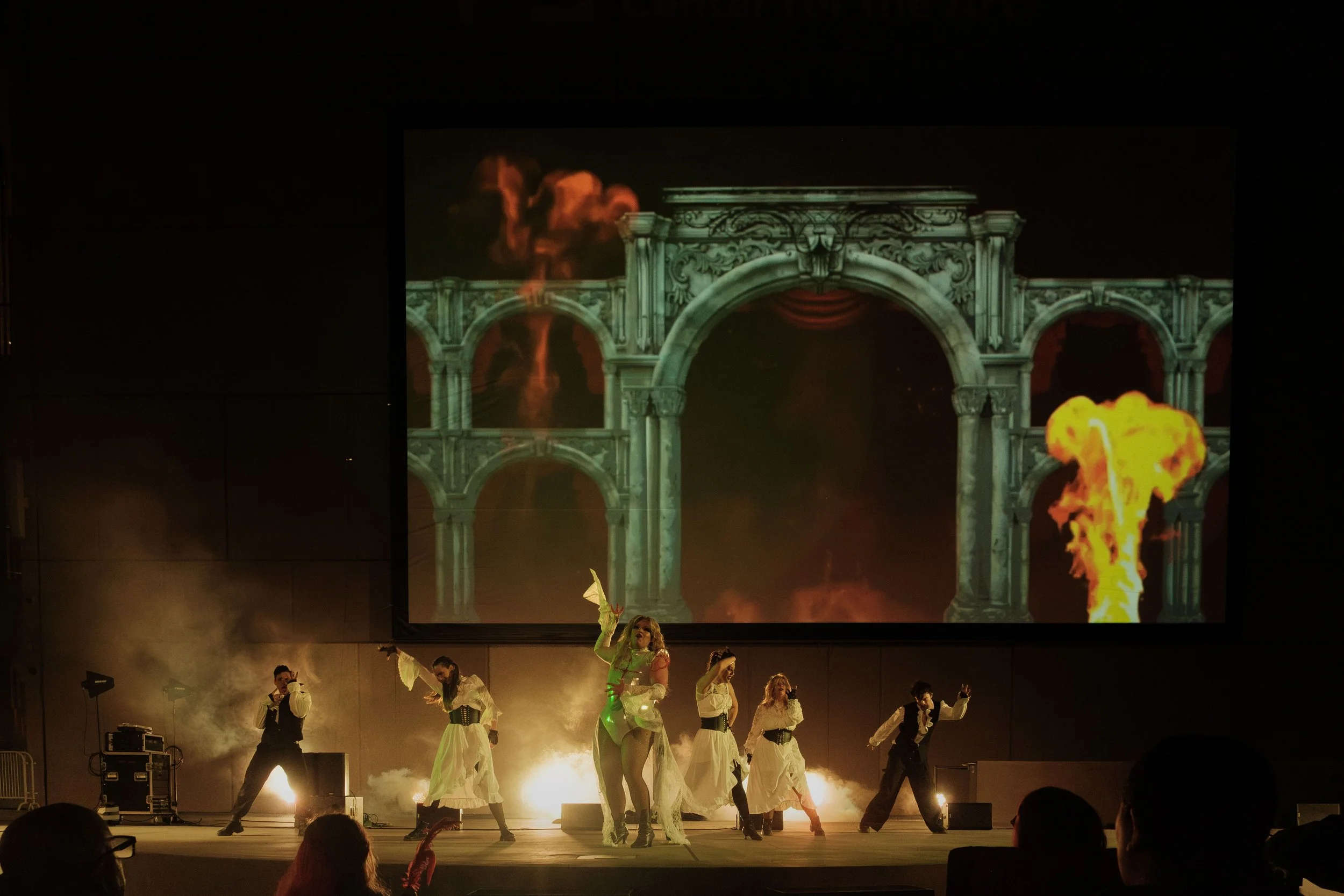 A theatrical performance with six performers on stage, some dressed in white costumes with black belts, others in dark suits. The stage has smoke effects and a large screen behind them displays a fantasy scene with ancient arches and flames. Broadway