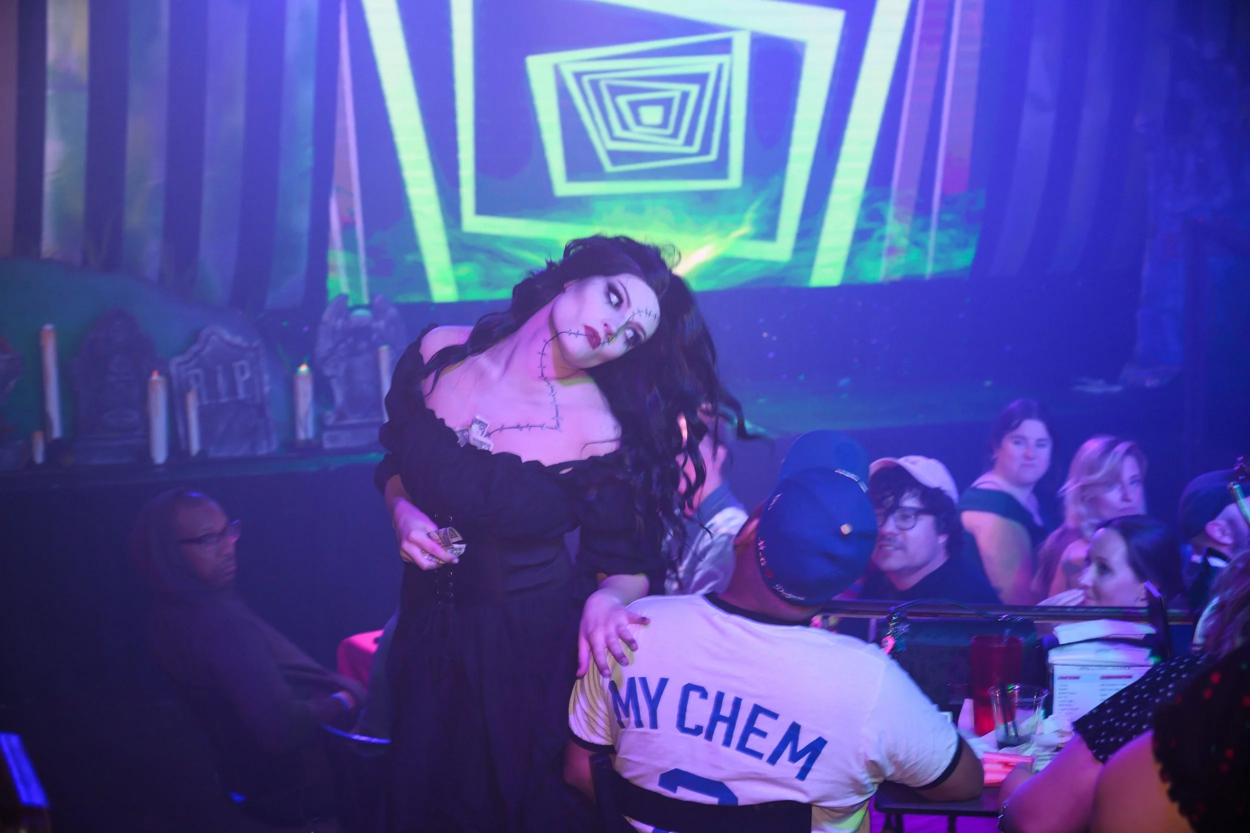 A woman dressed in a black costume with stitches painted on her face and chest, resembling a spooky character, is at a party or celebration. She is standing among seated and standing people at a table with drinks, in a setting with colorful, swirling, geometric digital displays on the screen behind her, creating a vibrant, festive atmosphere.