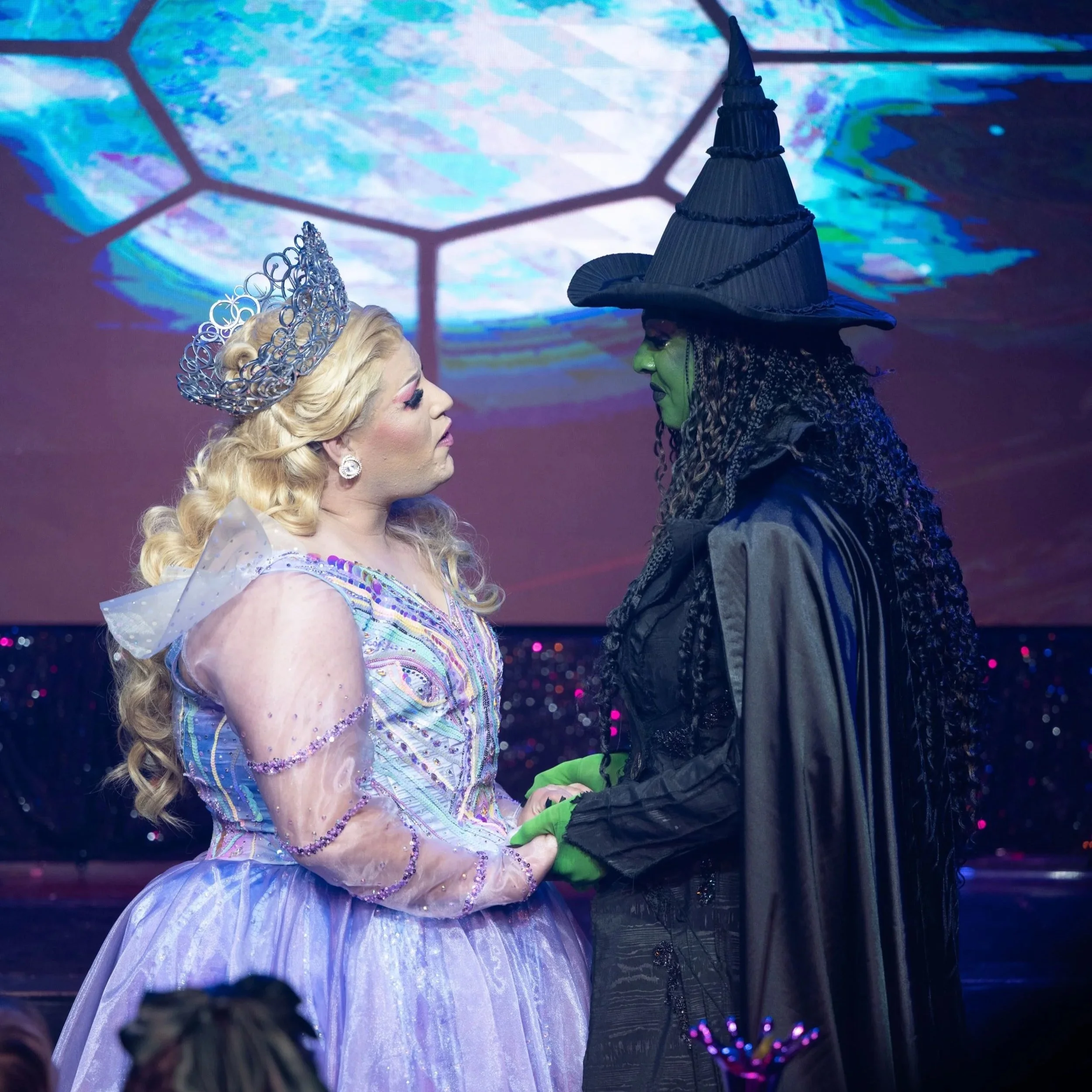 Two women dressed as characters from 'The Wizard of Oz' face each other closely on stage, holding hands. One is dressed as Glinda the Good Witch with a tiara and a lavender gown, and the other as the Wicked Witch of the West with a green face, black pointed hat, and dark cloak.