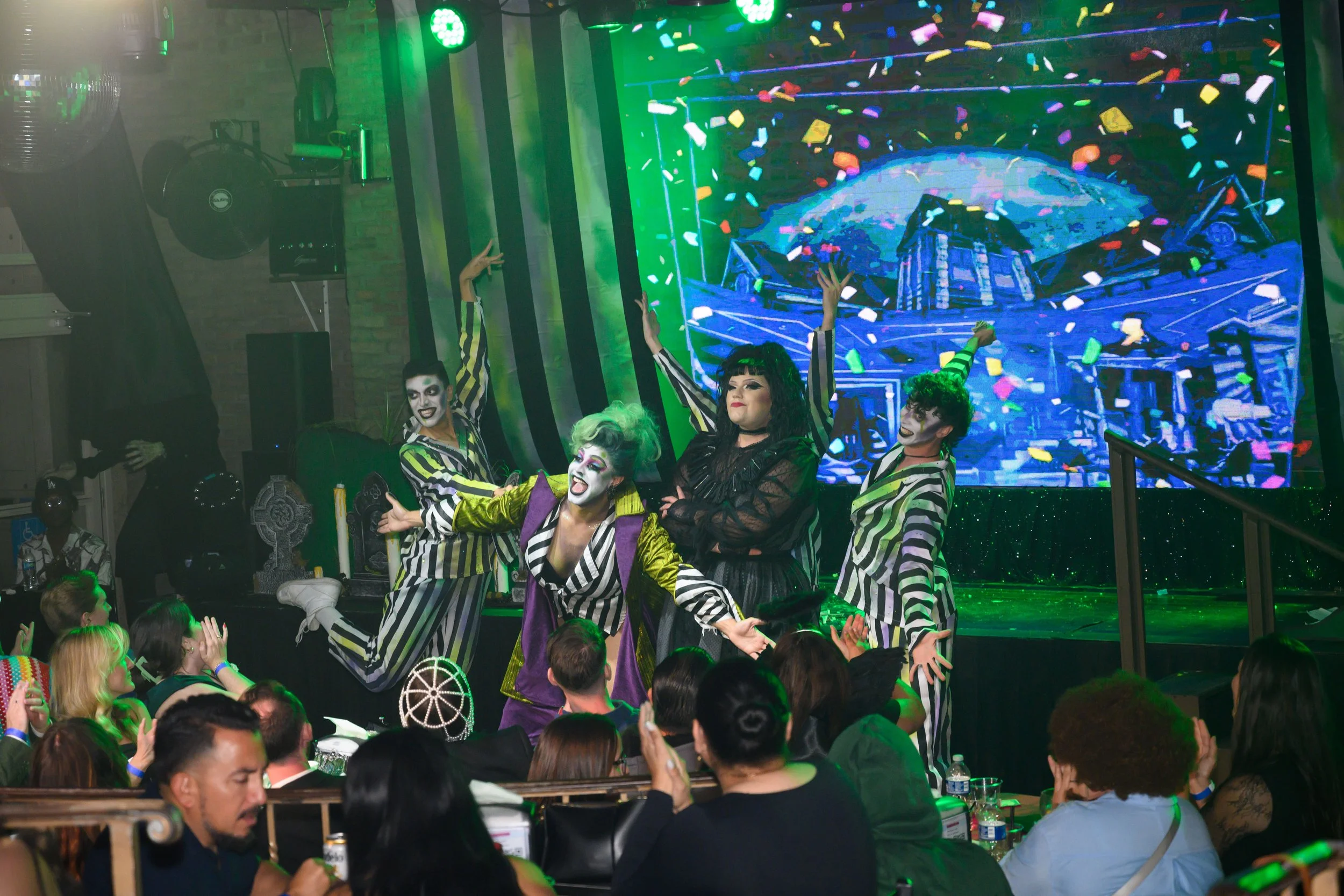 Performers dressed as clowns with theatrical makeup dance on a stage with a digital backdrop showing a cityscape and confetti, entertaining an audience at a lively event. Broadway Divas SCFTA