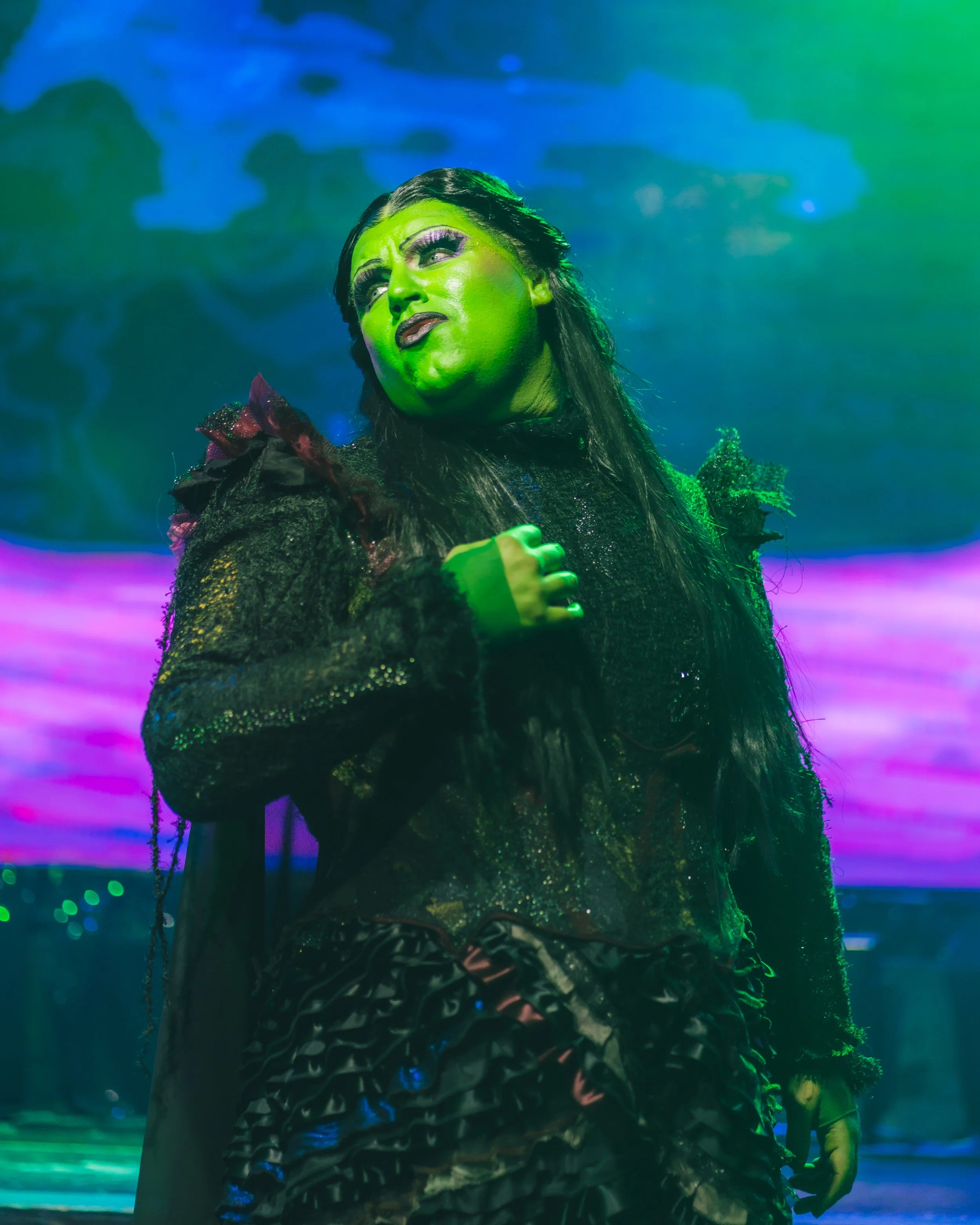 A person dressed in black with dramatic makeup and green face paint, wearing an elaborate costume, standing in front of a colorful, abstract background with neon lights. Wicked Elphaba Broadway Divas SCFTA Miss Clair Voyance