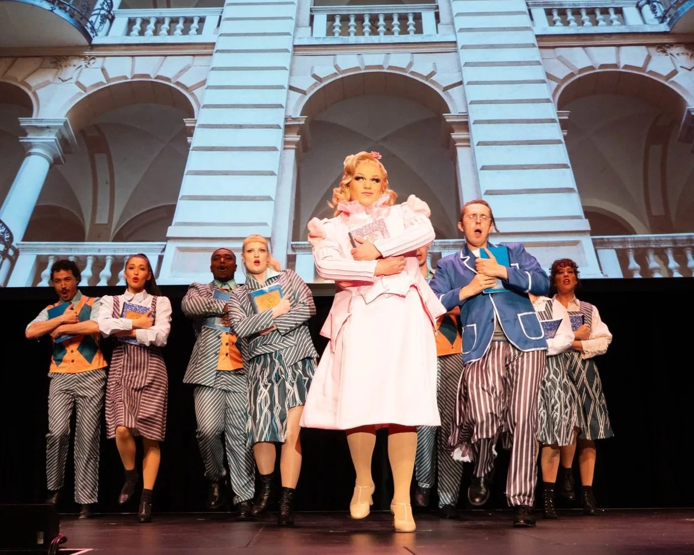Theatrical performance with actors on stage, lead actress in pink dress holding books, backdrop of grand building facade, group of actors in colorful costumes singing or performing. Twisted: For Good Broadway Divas SCFTA Miss Clair Voyance