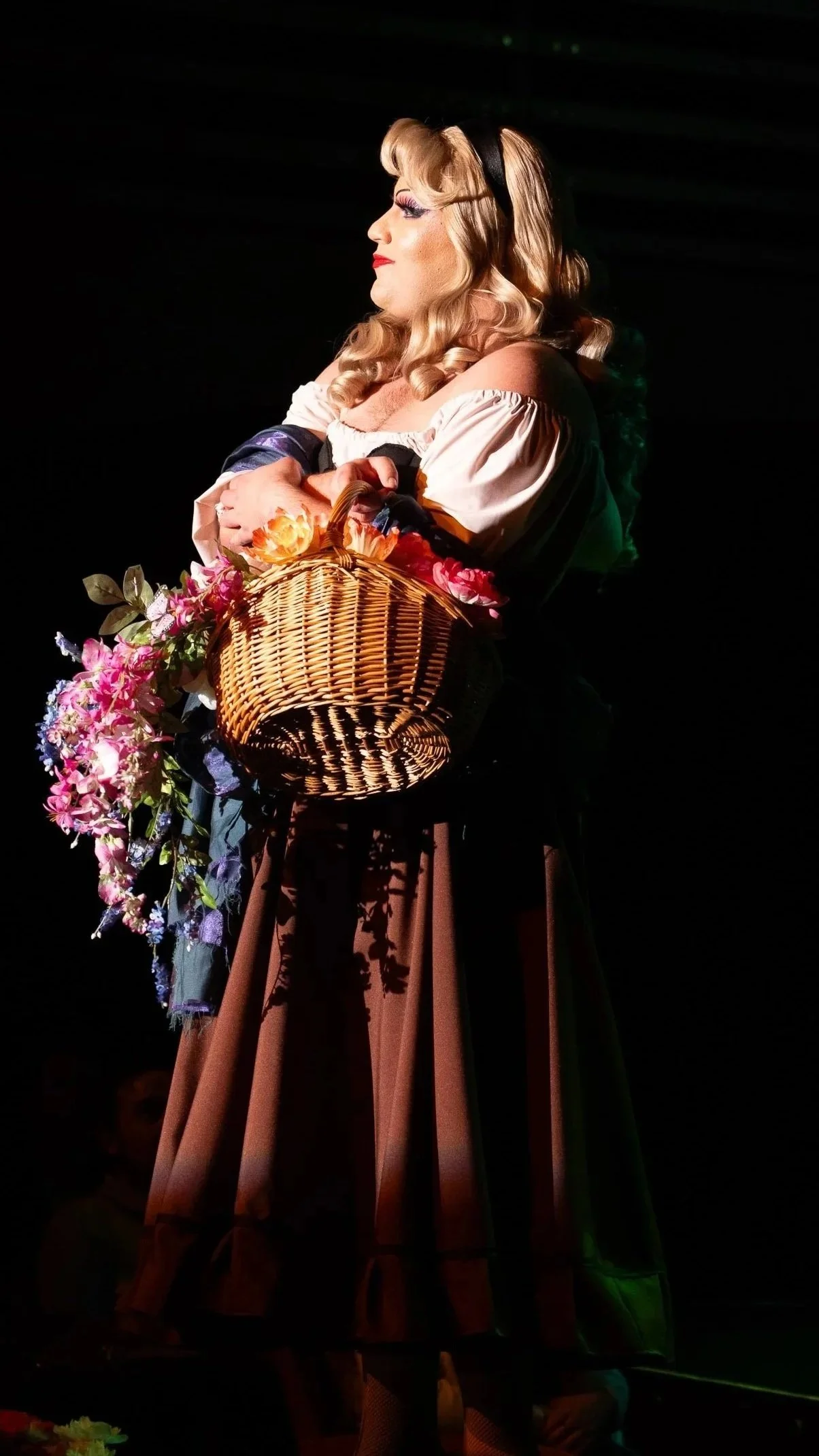 A woman dressed in vintage clothing with curled blonde hair, wearing makeup, holding a basket filled with colorful flowers, standing on stage with a dark background. Broadway Divas SCFTA