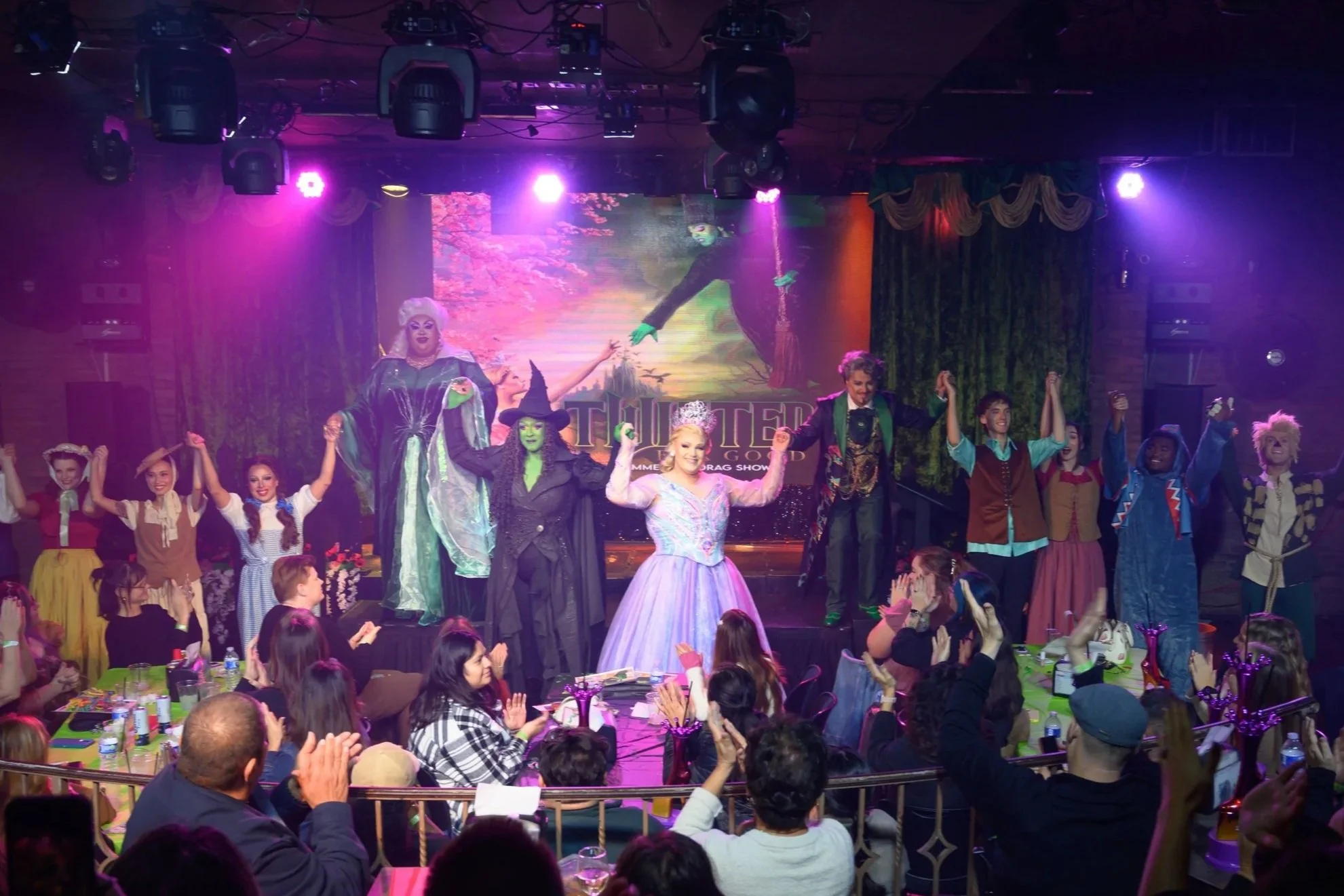 Stage scene from a theatrical performance featuring cast members dressed as witches, princesses, and fairy tale characters, with an audience at tables clapping and cheering in a dimly lit theater with purple lighting. Broadway Divas SCFTA
