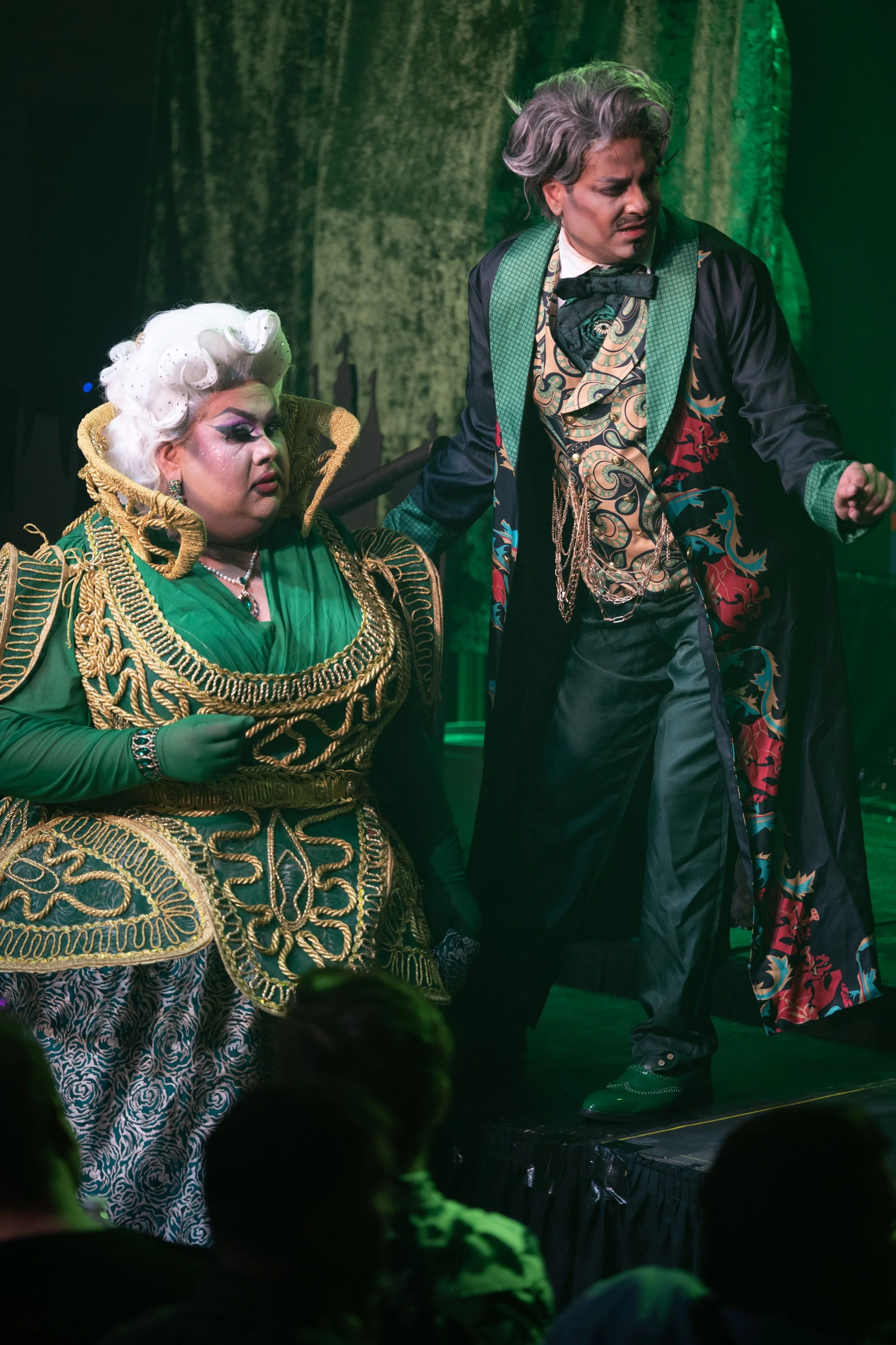 Two performers in elaborate costumes on stage during a theatrical performance, with audience members visible in the foreground. Broadway Divas SCFTA