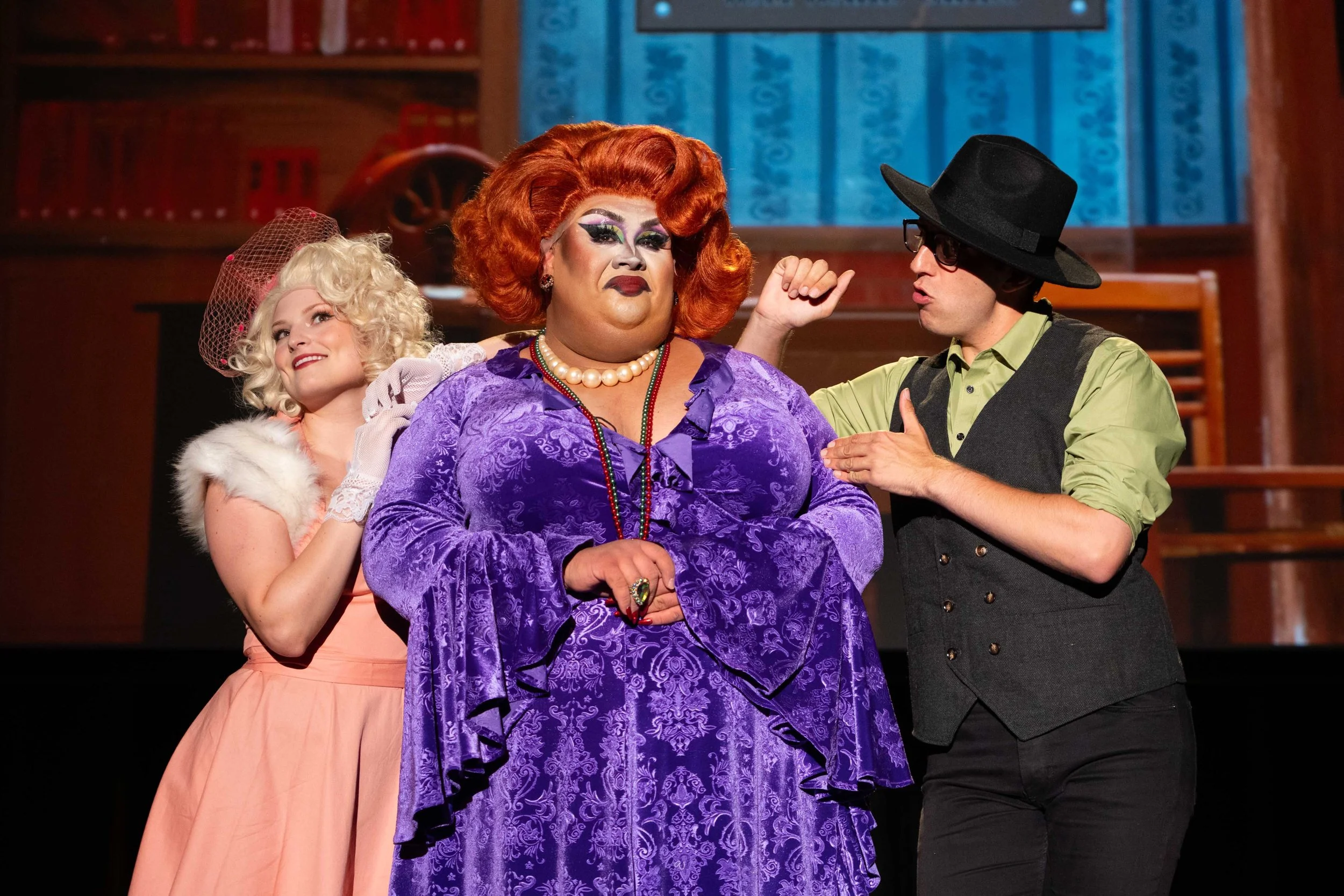 Three actors on stage in colorful costumes, one with red hair in a purple dress, another with blonde curly hair in a pink dress with a fur stole and hat, and a man in a black hat, glasses, and vest, engaging in a theatrical scene. Broadway Divas SCFT