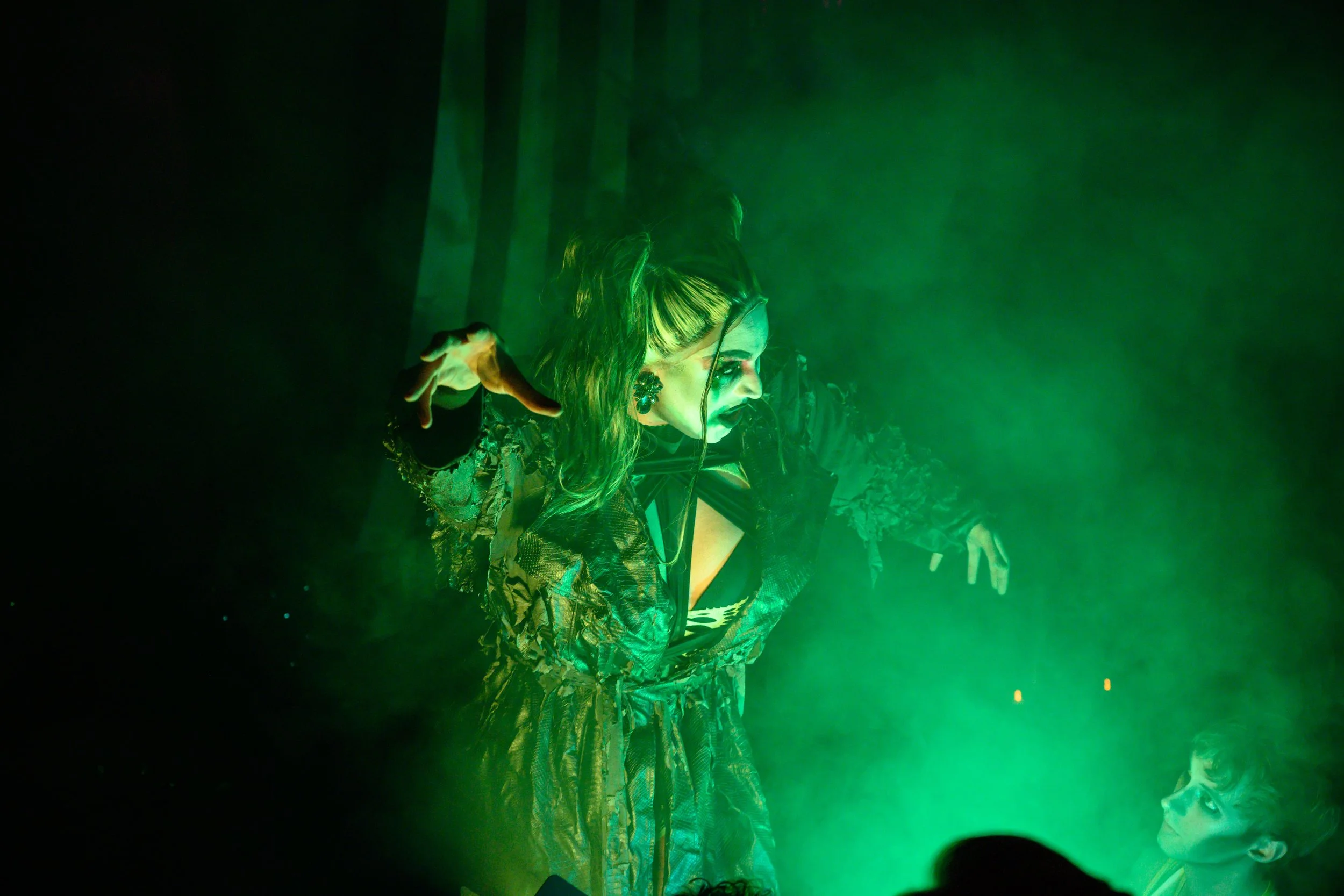 Performer with green lighting, wearing a patterned dress, makeup, and glasses, gesturing with one hand, with another person in glasses looking up at them. Broadway Divas SCFTA