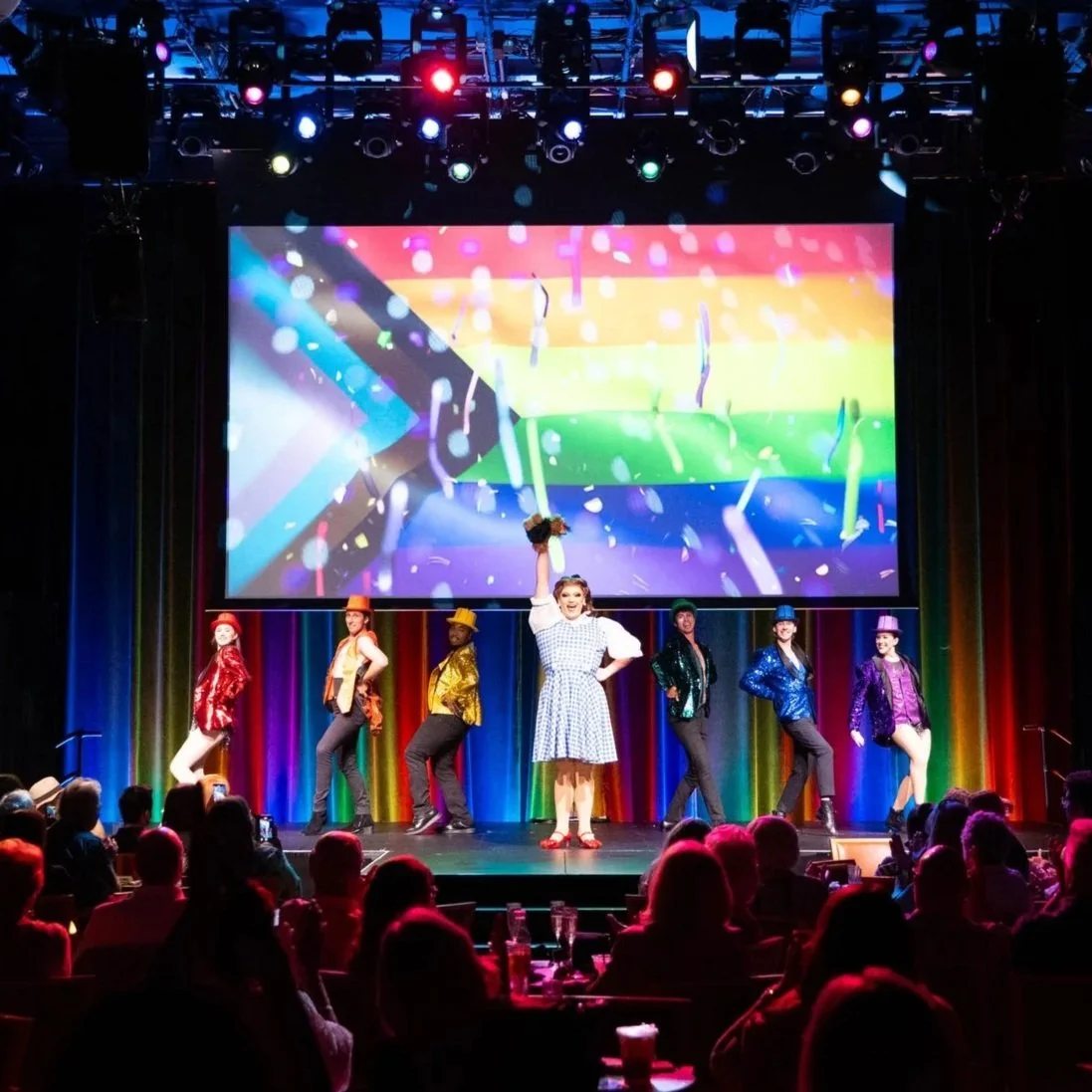 A musical or theatrical performance on stage with a group of six performers dressed in colorful costumes. One woman in the center holds a bouquet and raises her arm. Broadway Divas SCFTA Miss Clair Voyance