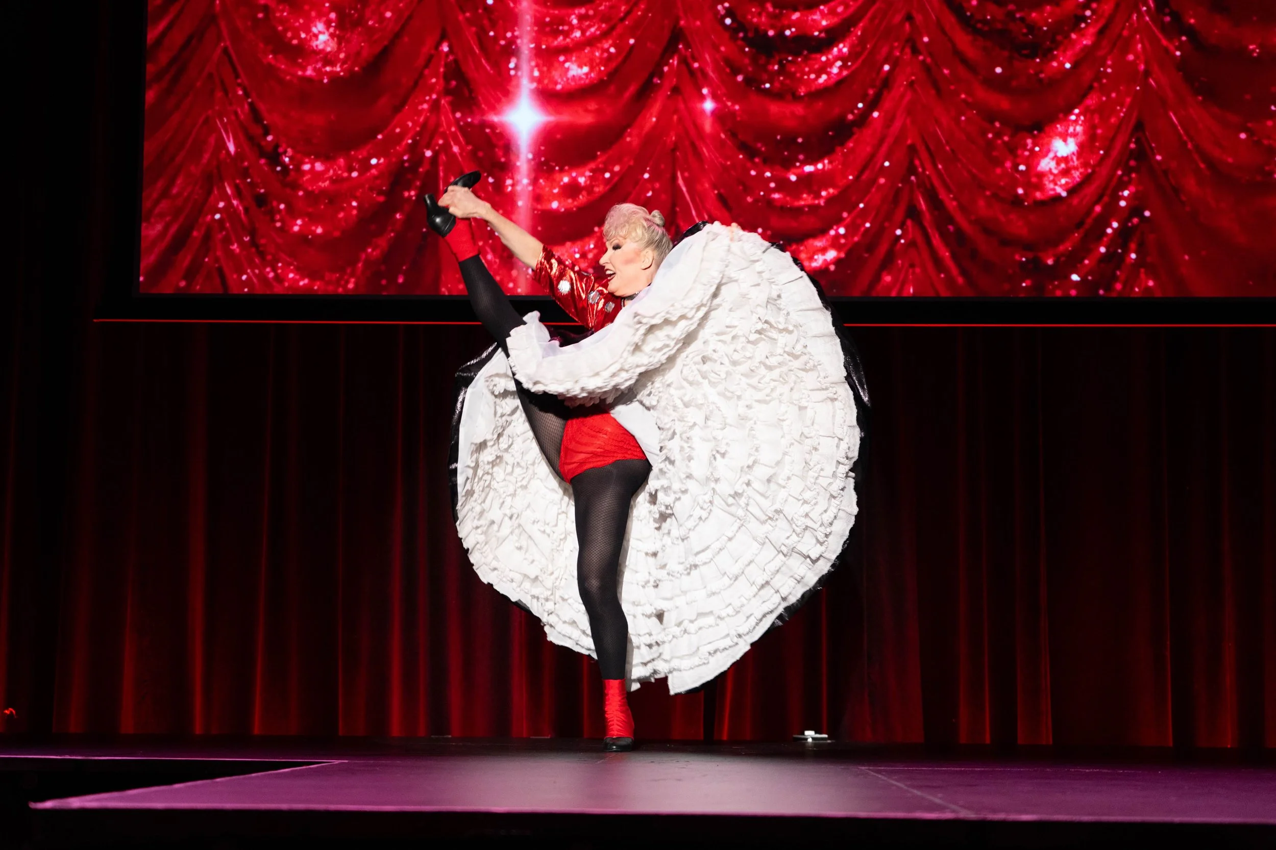 A performer on stage in a red outfit, fishnet stockings, and high heels, holding one leg up while wearing a large white frilly costume, with a red curtain backdrop and a digital display showing a red curtain with sparkles. Broadway Divas SCFTA