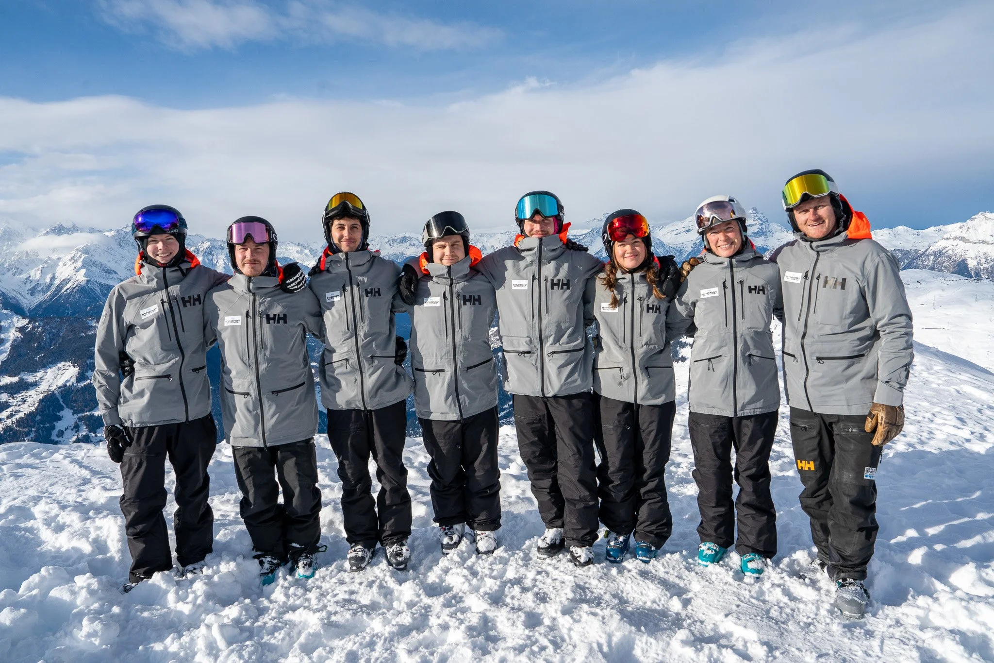 Group of nine people in winter gear standing on snow-covered ground with snowy mountains and blue sky in the background.