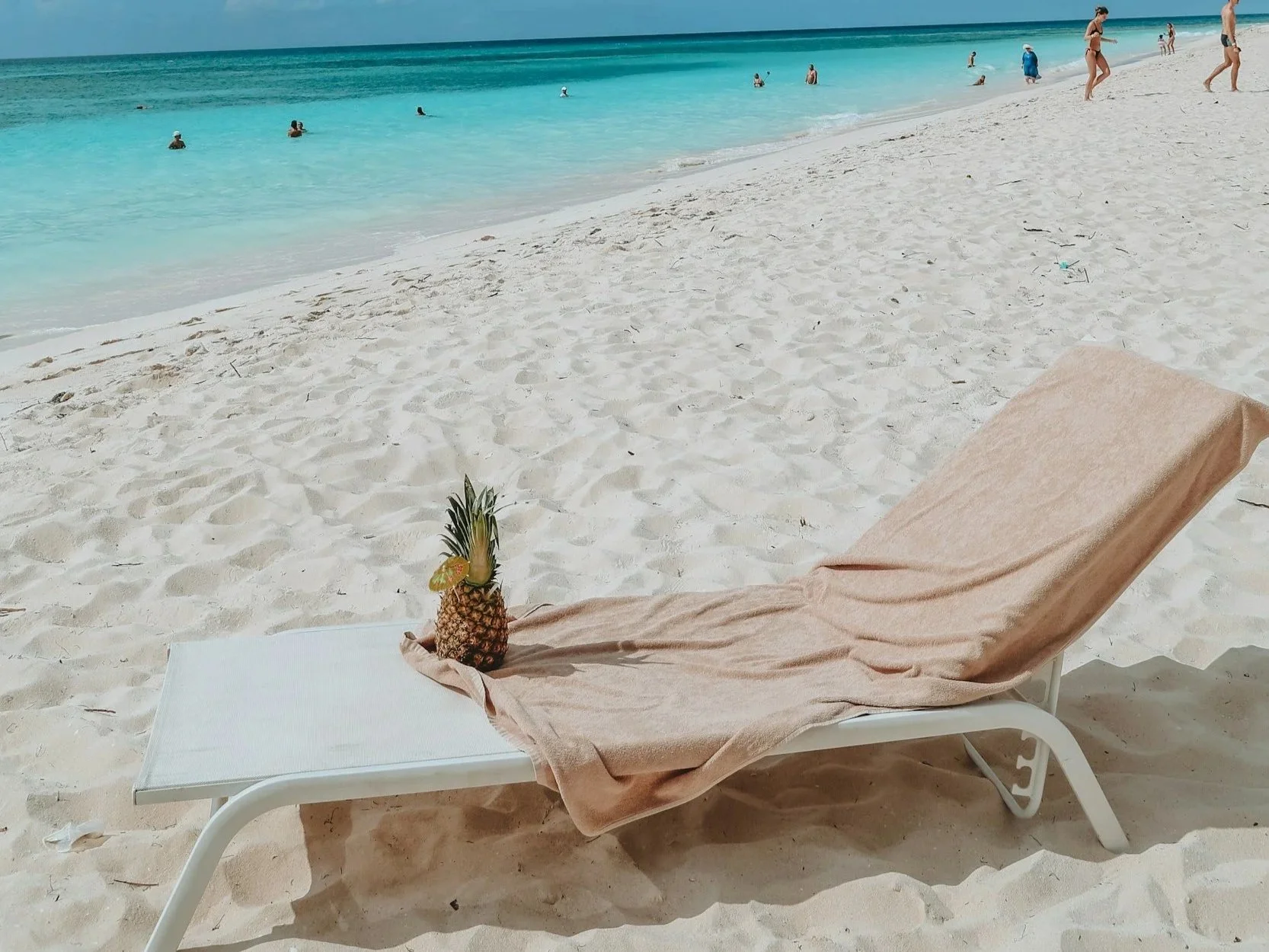 A beach scene with a lounge chair and a pineapple with a straw and umbrella on it. The beach has white sand and turquoise water, with people swimming and walking along the shoreline.