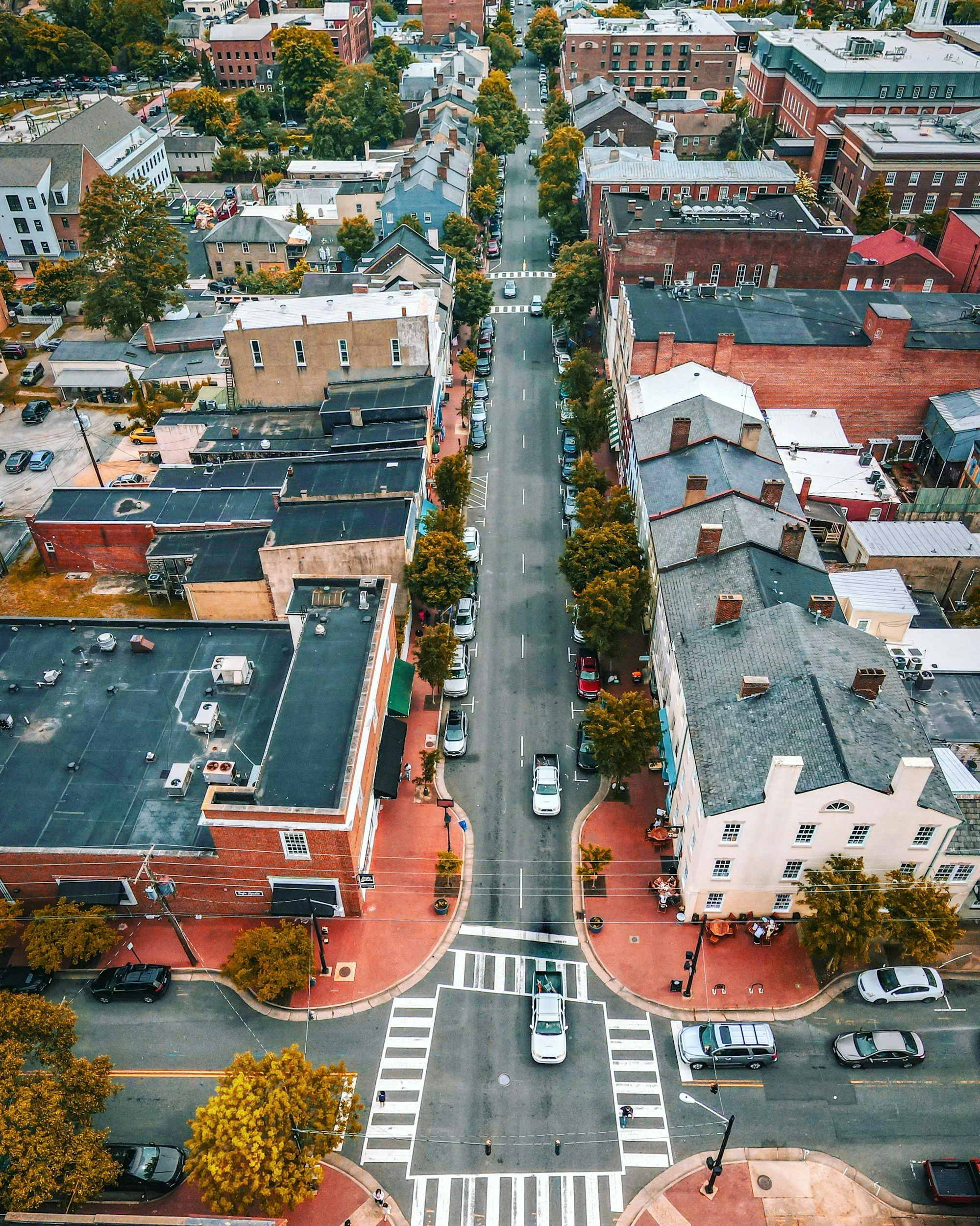 An aerial view of a downtown city street with parked cars, trees lining the sidewalks, and various buildings on both sides, including brick and commercial structures.
