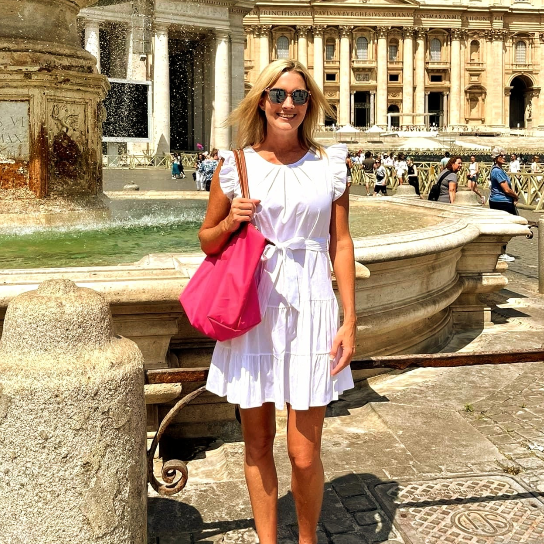 A smiling woman with blonde hair wearing sunglasses and a white dress, holding a pink bag, standing in front of a fountain at a historic Roman-style building.