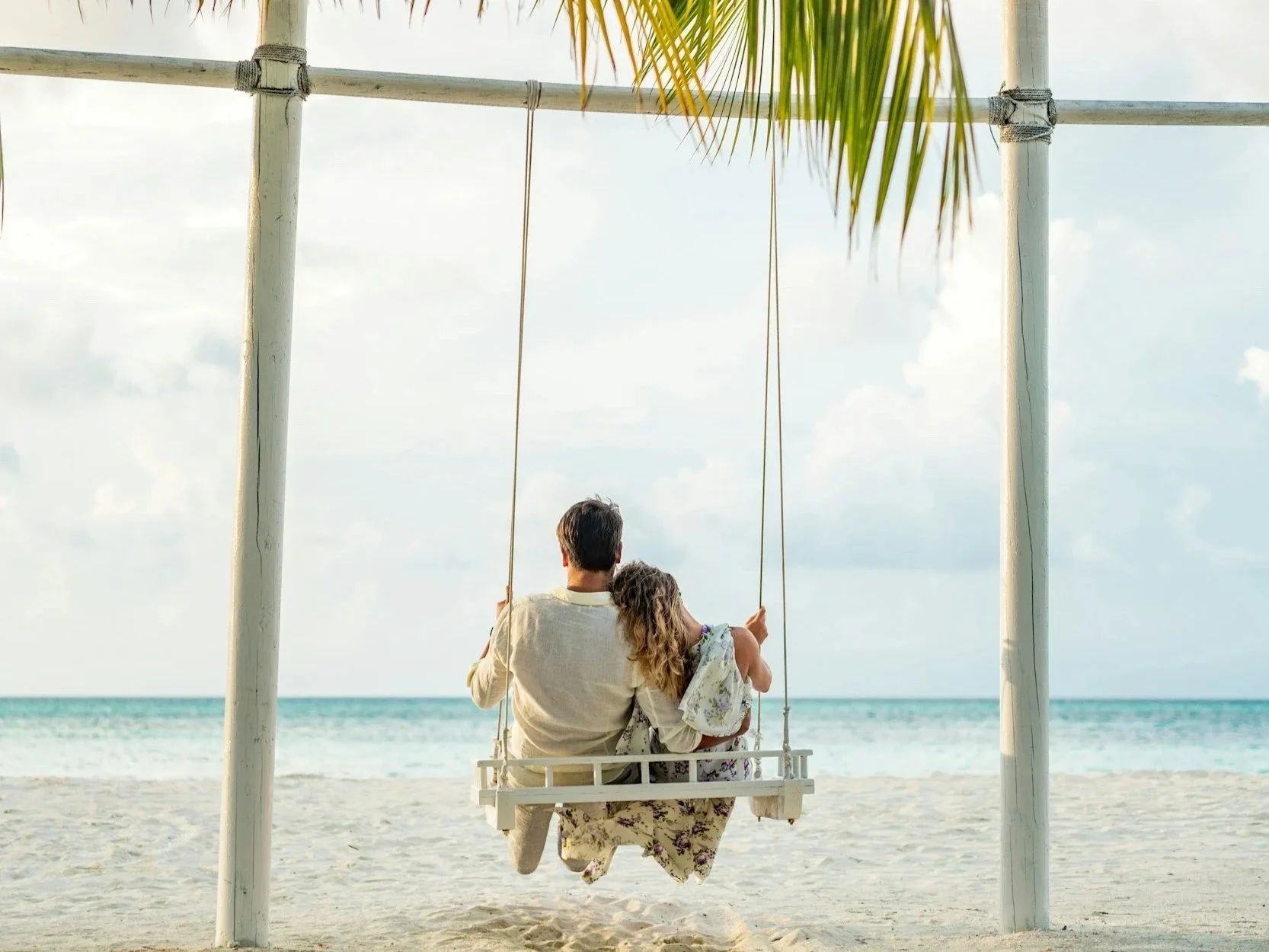 A man and a girl sitting together on a swing at the beach, facing the ocean, with a palm tree leaf hanging overhead.