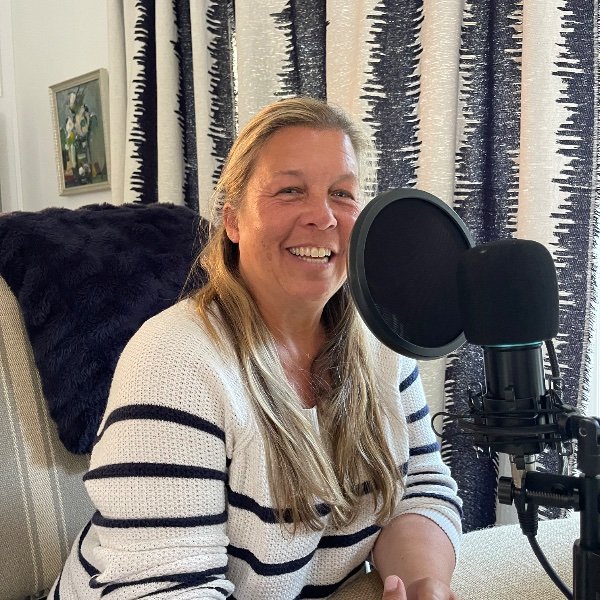 A woman with long blonde hair sitting at a table with a microphone in front of her, smiling at the camera in a room with patterned curtains and a black pillow behind her.
