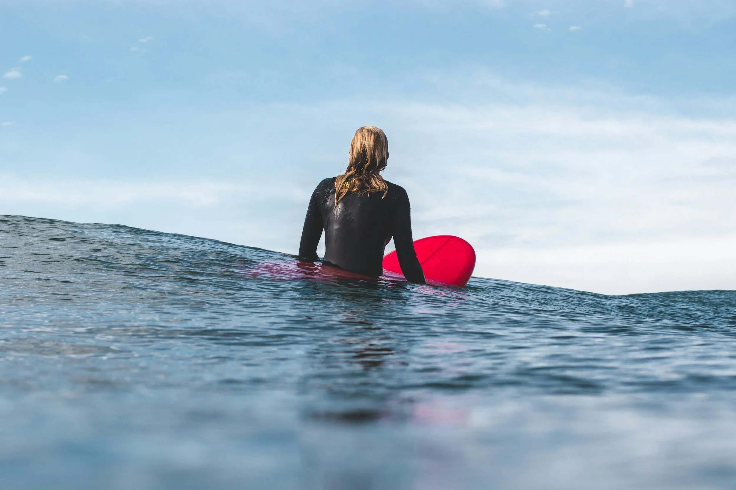 Person in wetsuit holding a red surfboard sitting on a wave in the ocean, looking at the sky.