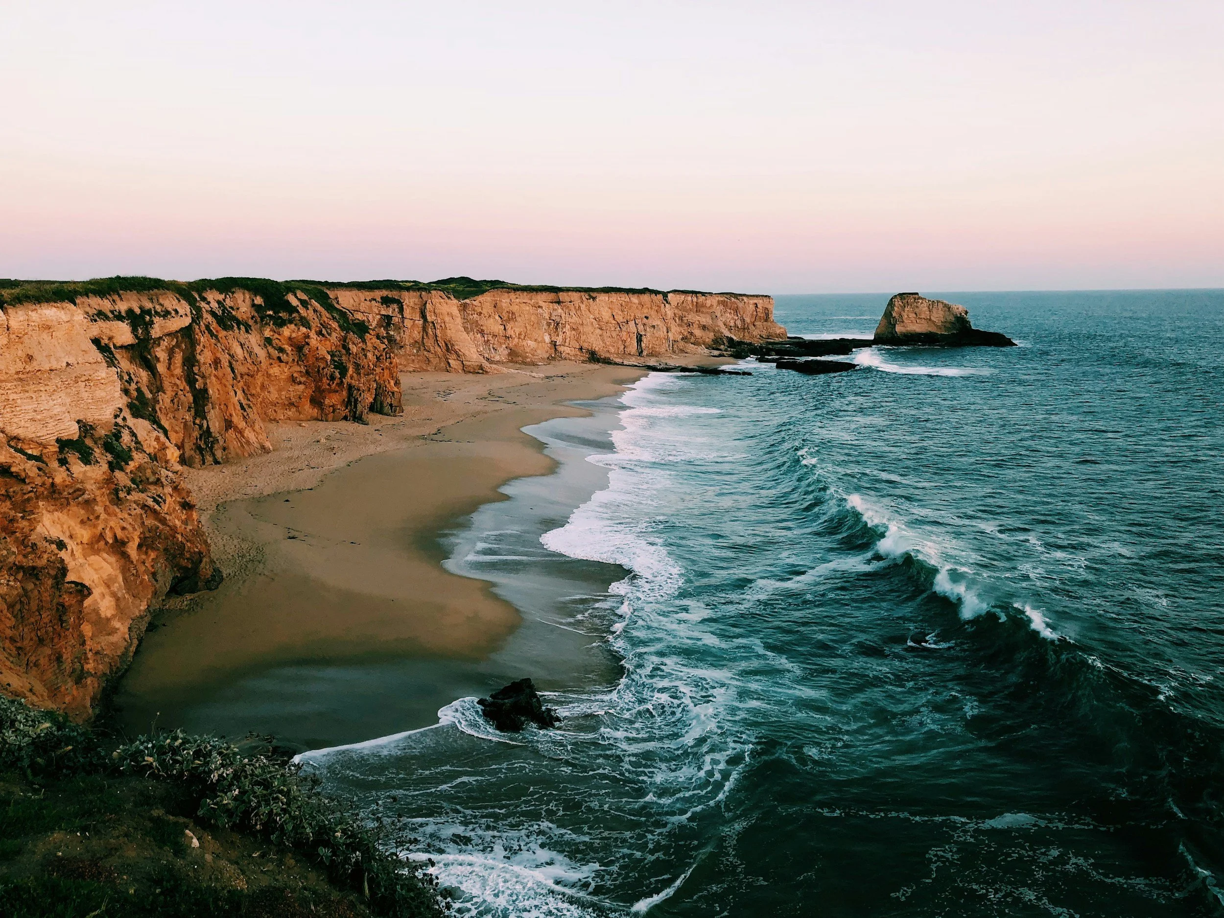 Cliffs overlooking a sandy beach and ocean with waves crashing, under a pastel sky.