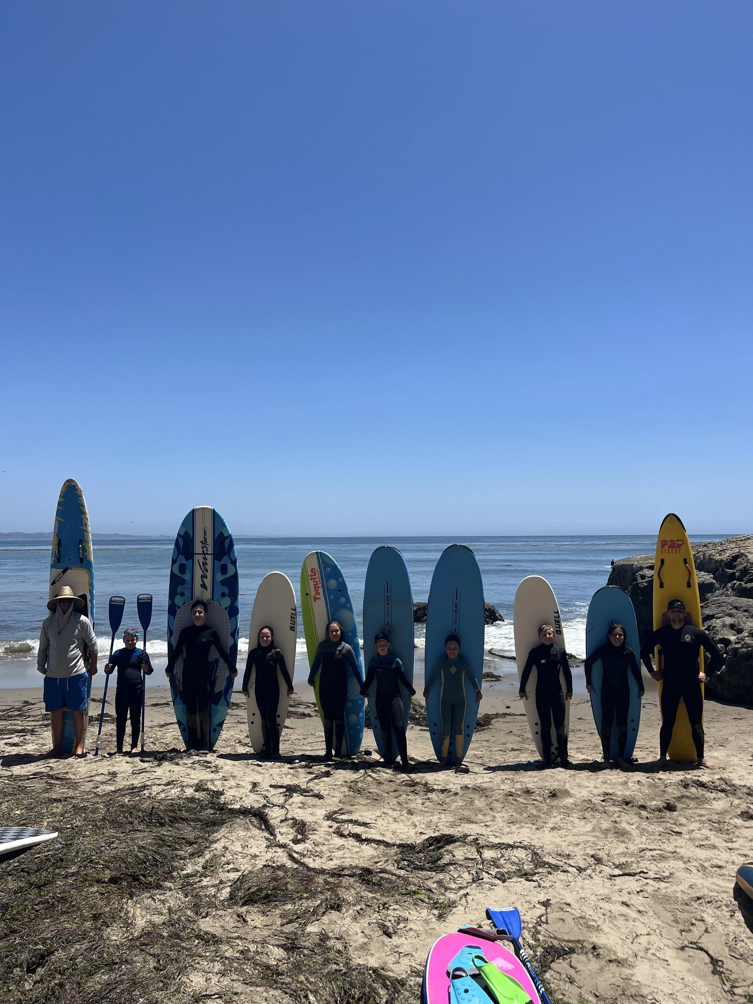 A group of ten people, including children and adults, standing on the beach with surfboards, paddleboards, and a kayak, ready for a surfing lesson from Ocean X Acents Training.