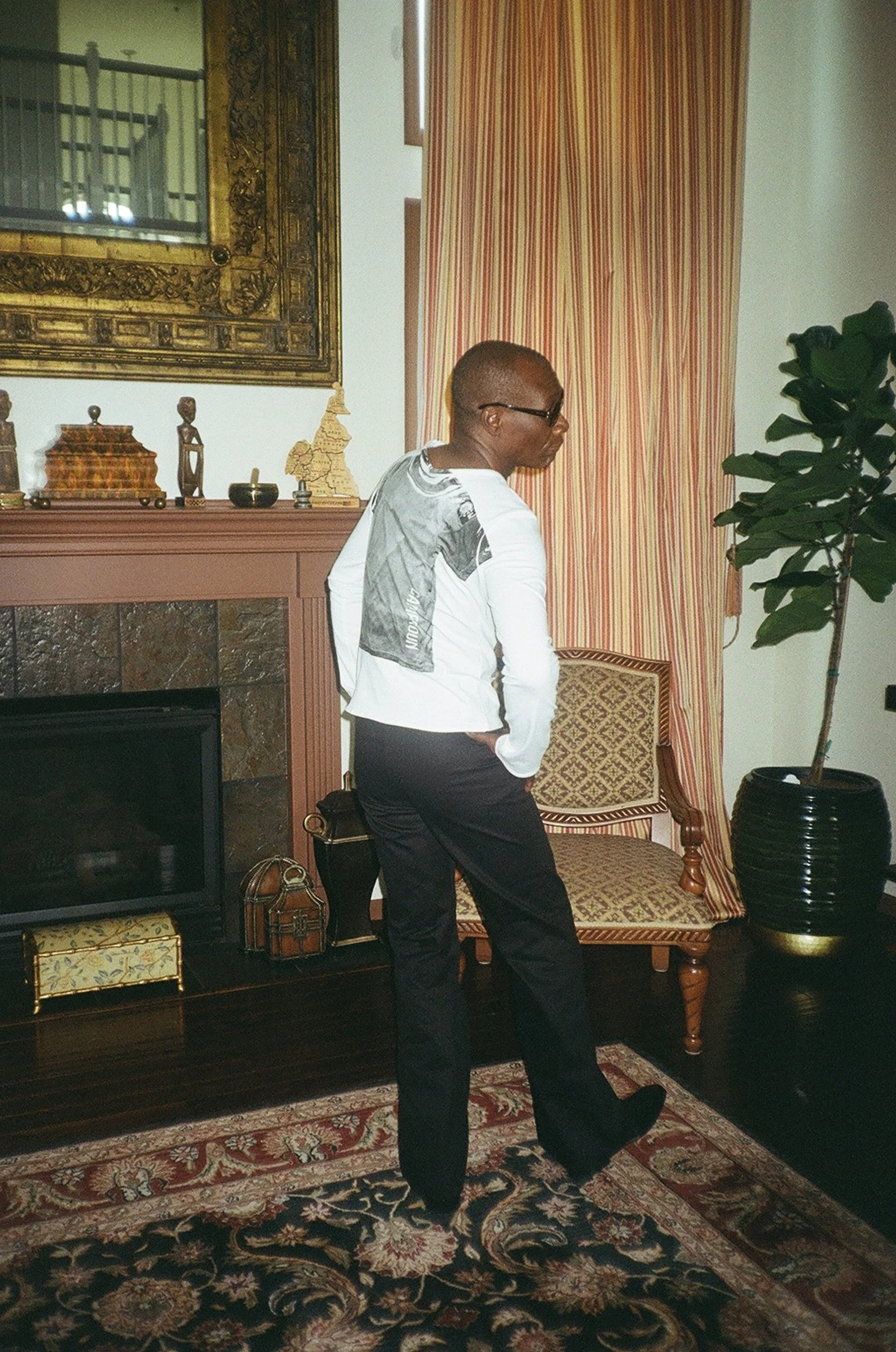 A man with his back facing the camera. Posing with his right leg up wearing a white long sleeve and black flared trousers.