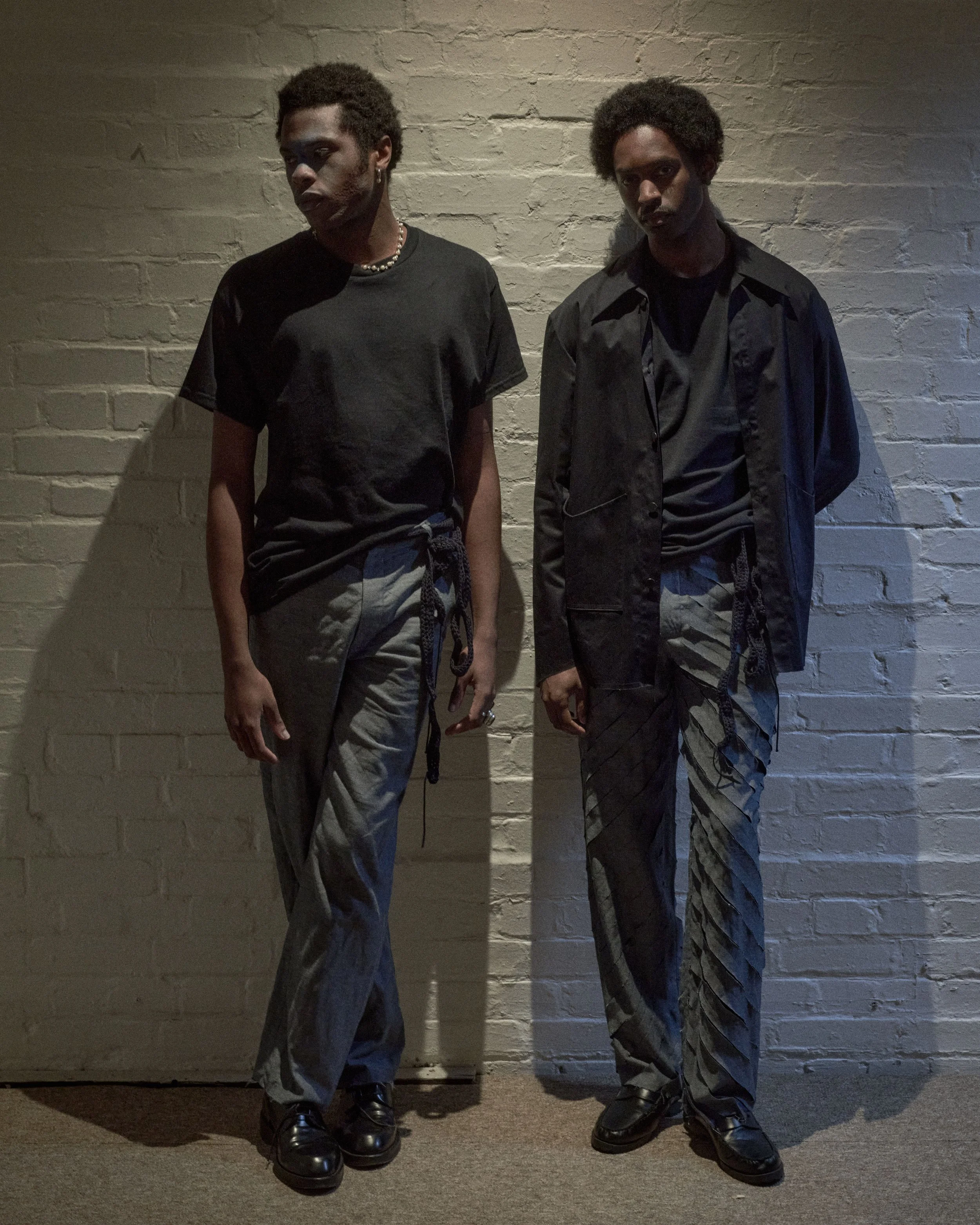 Two young men with dark curly hair leaning against a white brick wall, dressed in black outfits with patterned pants and black shoes, posing in a dimly lit room.