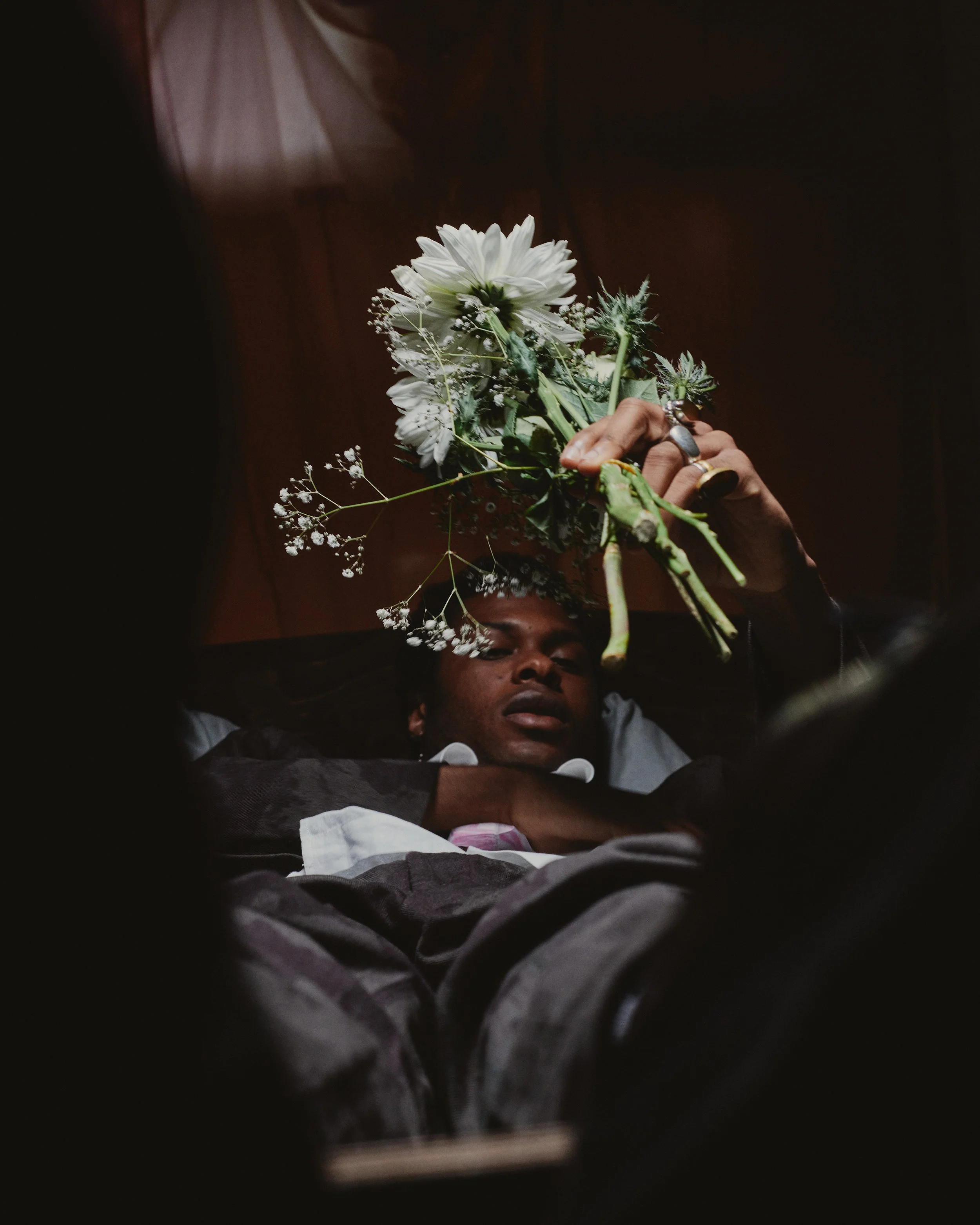 A person lying down holding a bouquet of white flowers over their forehead, with a dark background.