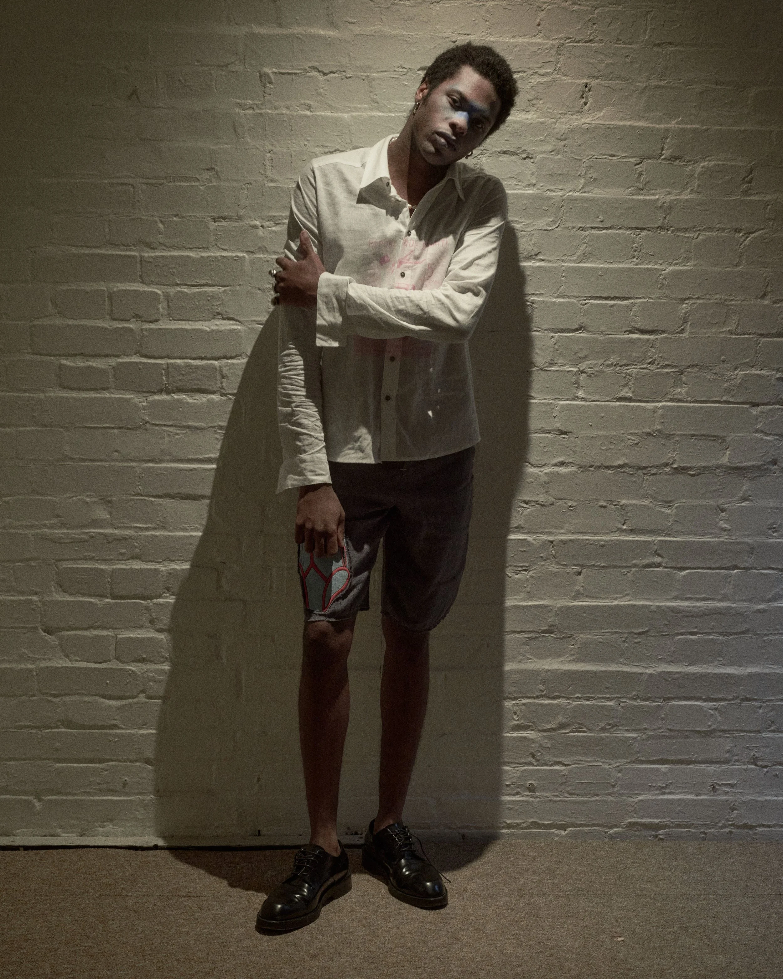 A young man standing against a white brick wall, wearing a white button-up shirt, shorts, and black shoes, holding a phone in his left hand.