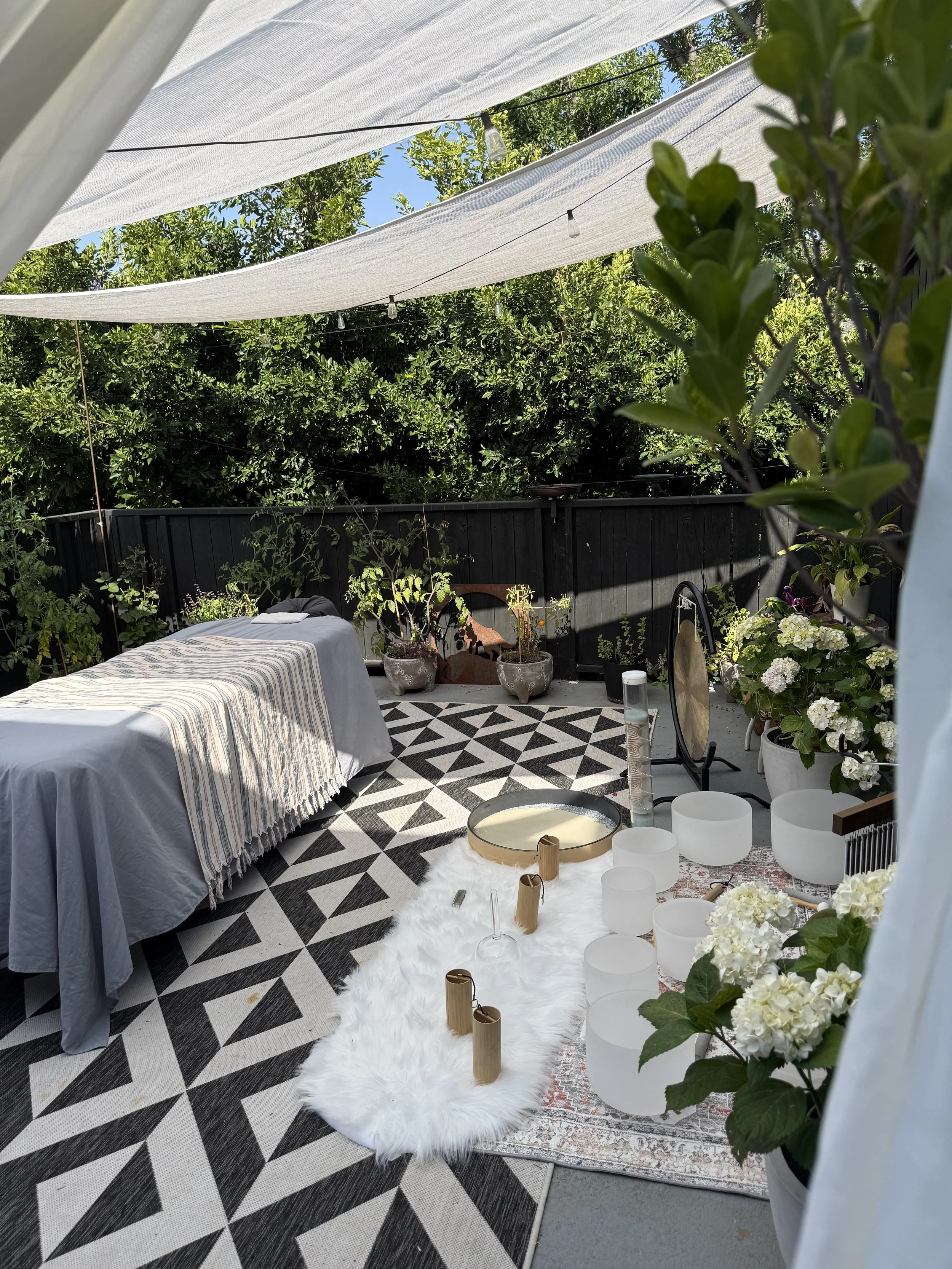 Outdoor patio decorated with white drapes and string lights, featuring a table with a striped tablecloth, potted plants, and a fluffy white rug with metal and ceramic decor items. Sound bath instruments.