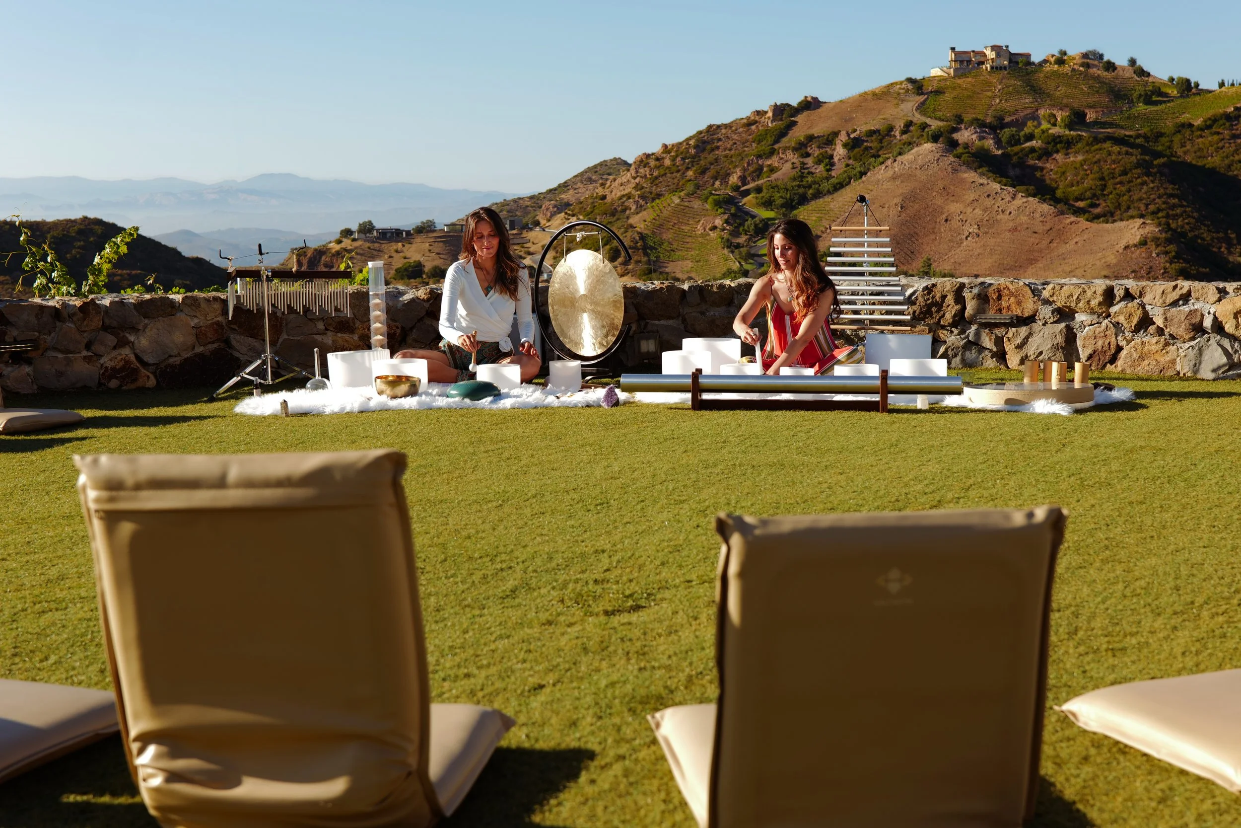 Two women sitting on a blanket with singing bowls and a gong, outdoors on a grassy area, with hills and houses in the background. Sound bath instruments.