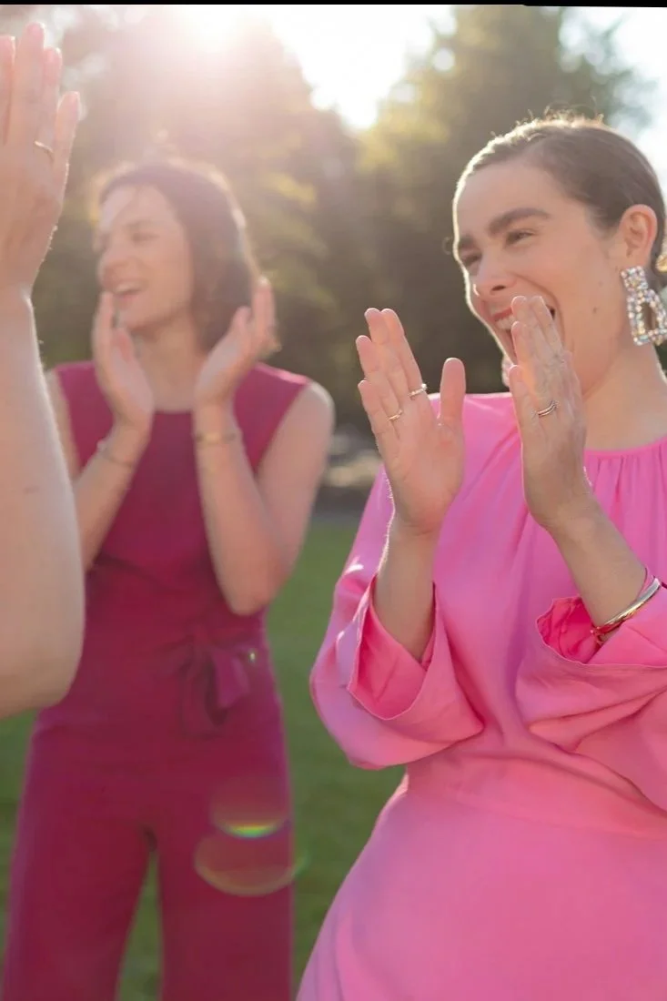 Drei Frauen lachen und klatschen draußen in der Sonne, bei einem Treffen im Park, bei angenehmem Wetter, elegante Kleidung, fröhliche Stimmung.