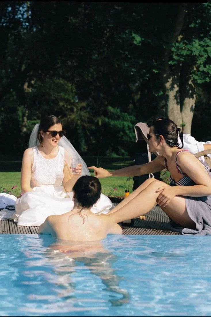 Eine Gruppe von Frauen und einem Mann entspannt am Pool an einem sonnigen Tag im Freien, umgeben von grünen Bäumen, einige sitzen auf der Poolkante, eine trägt eine Brautjungfernkleid, andere schwimmen im Pool.