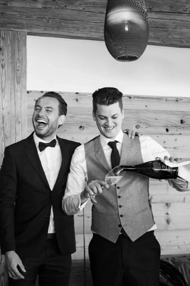 Vier Männer in formeller Kleidung, einige lachen und trinken Sekt, während sie in einem Raum mit Holzvertäfelung sind.