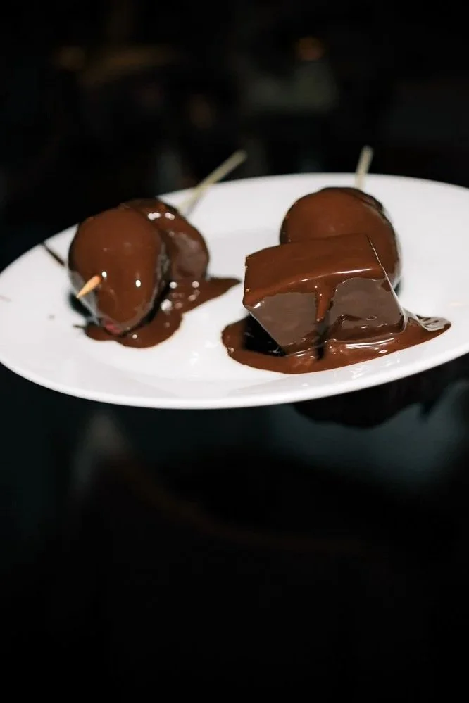 Ein Teller mit drei Schokoladen- oder Dessertspeisen, die mit Schokoladensauce überzogen sind, gehalten von einer Person, die eine Bluse mit blauen Mustern trägt.